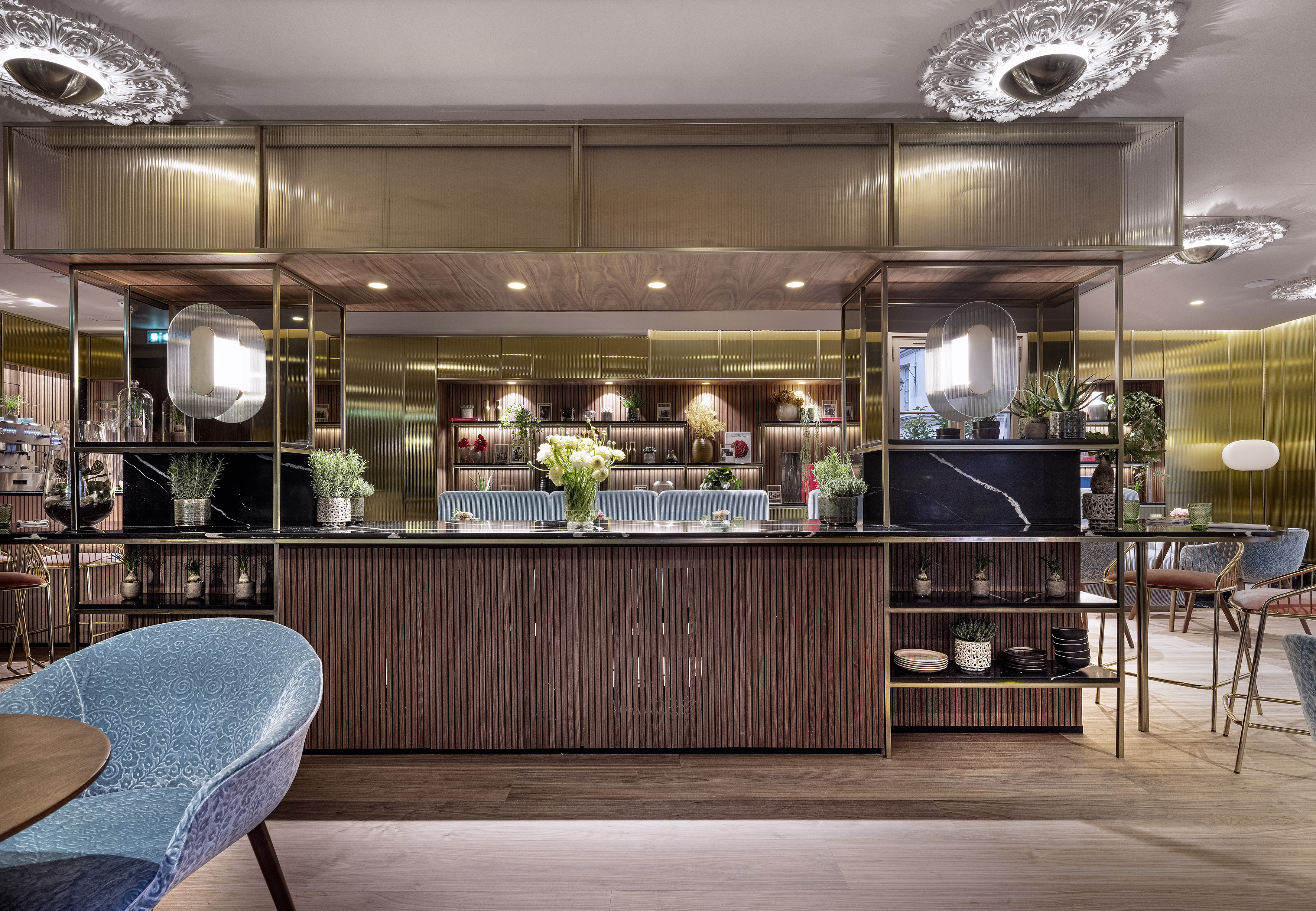 Modern hotel bar with wooden slatted counter, brass details, and lush plants.