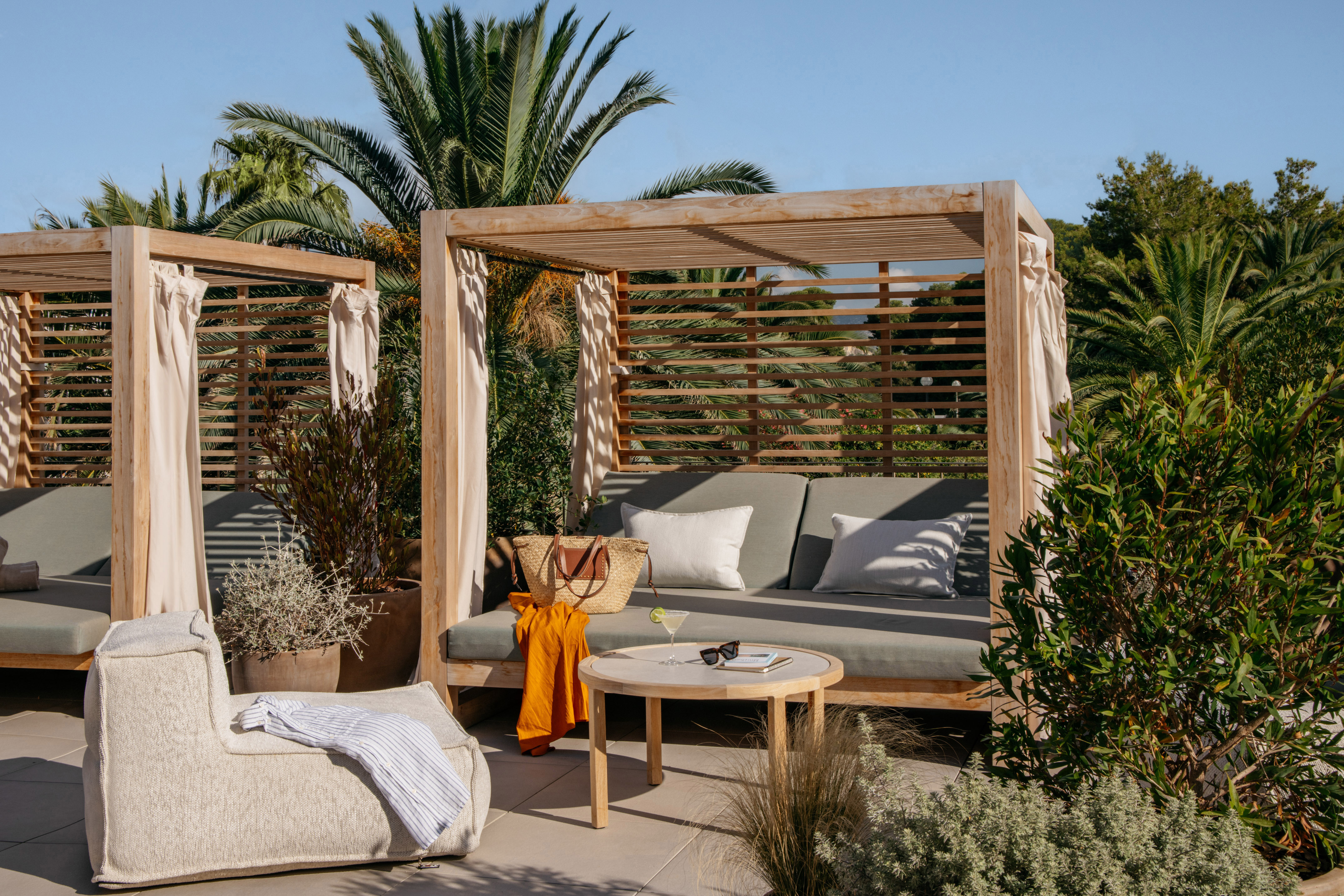 Sunny outdoor resort cabana with wooden daybed, cocktail, and tropical plants.