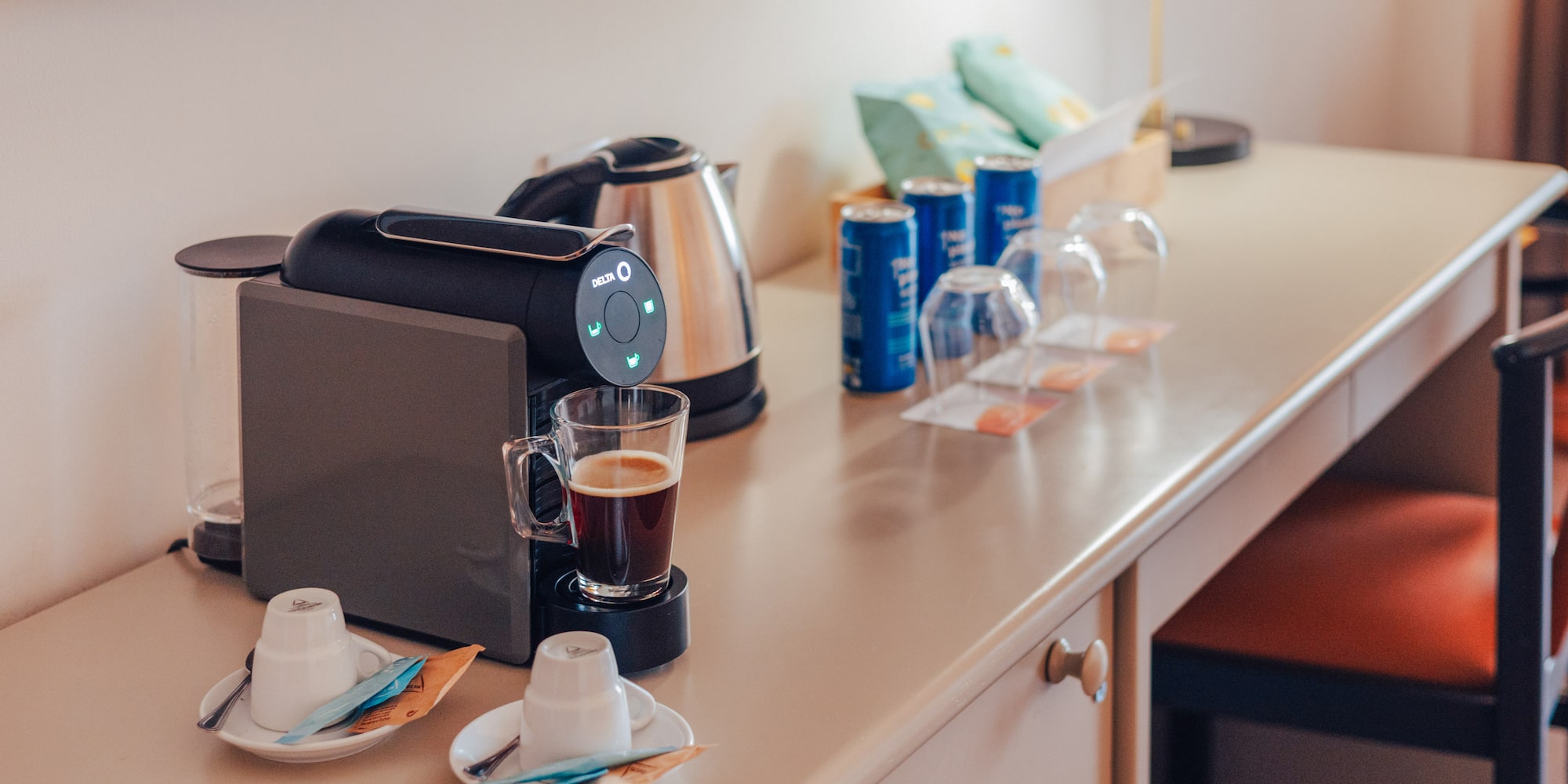Hotel room desk with coffee machine, kettle, coffee, cups, and snacks.