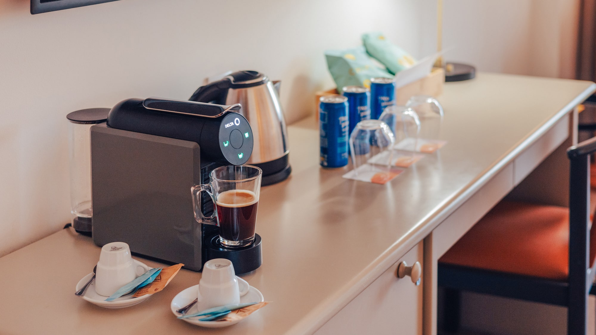 Hotel room desk with coffee machine, kettle, coffee, cups, and snacks.