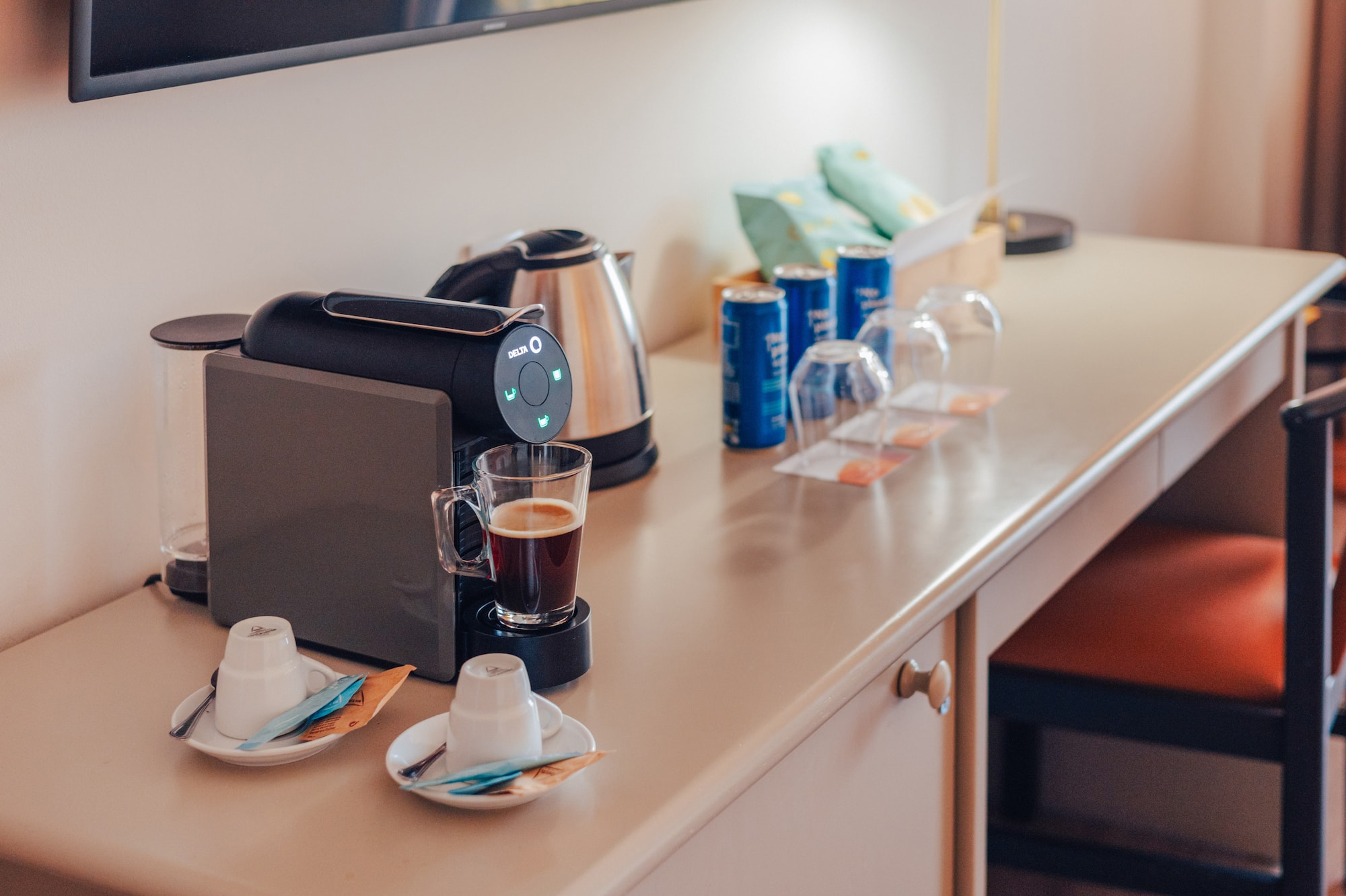 Hotel room desk with coffee machine, kettle, coffee, cups, and snacks.