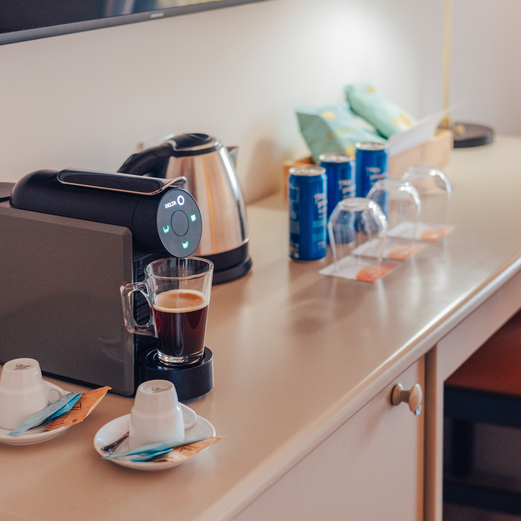 Hotel room desk with coffee machine, kettle, coffee, cups, and snacks.