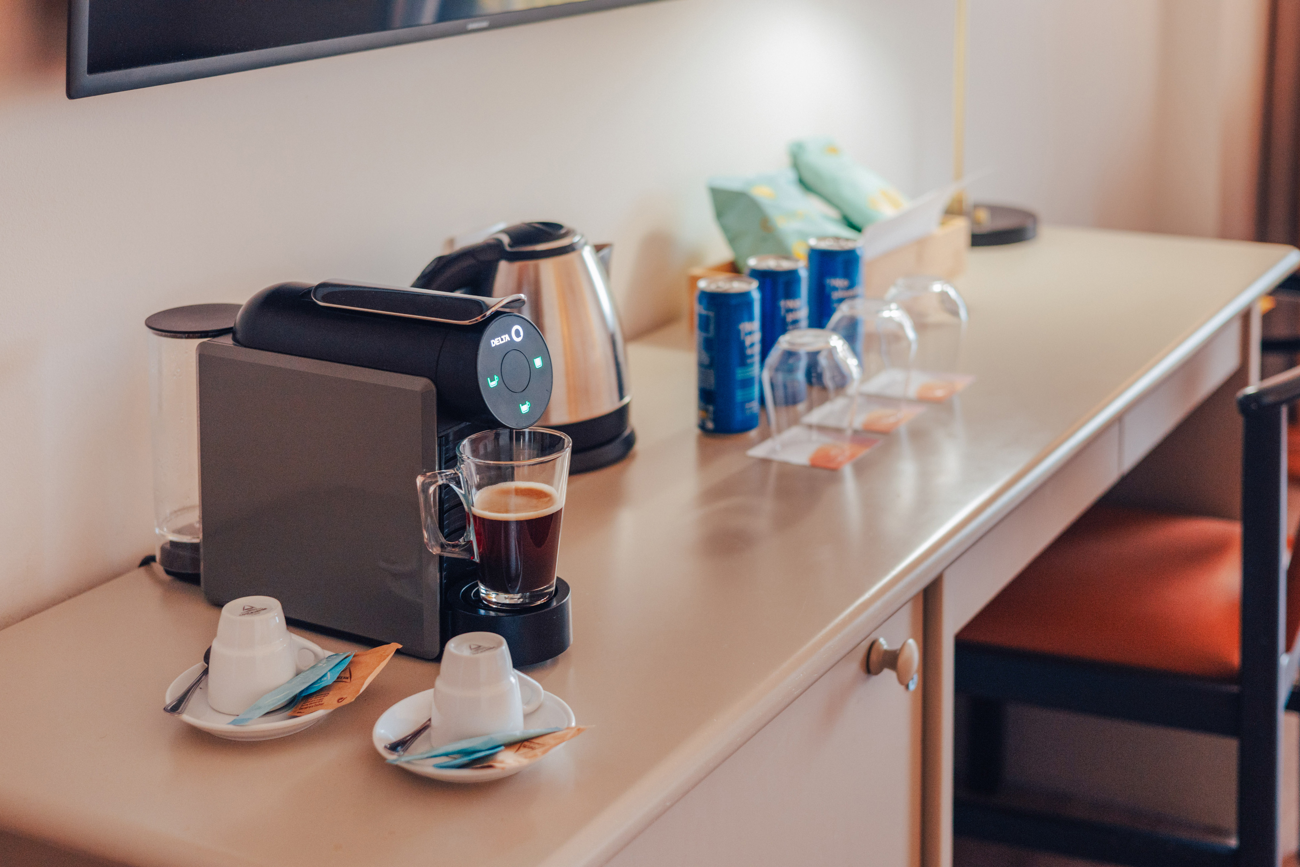 Hotel room desk with coffee machine, kettle, coffee, cups, and snacks.