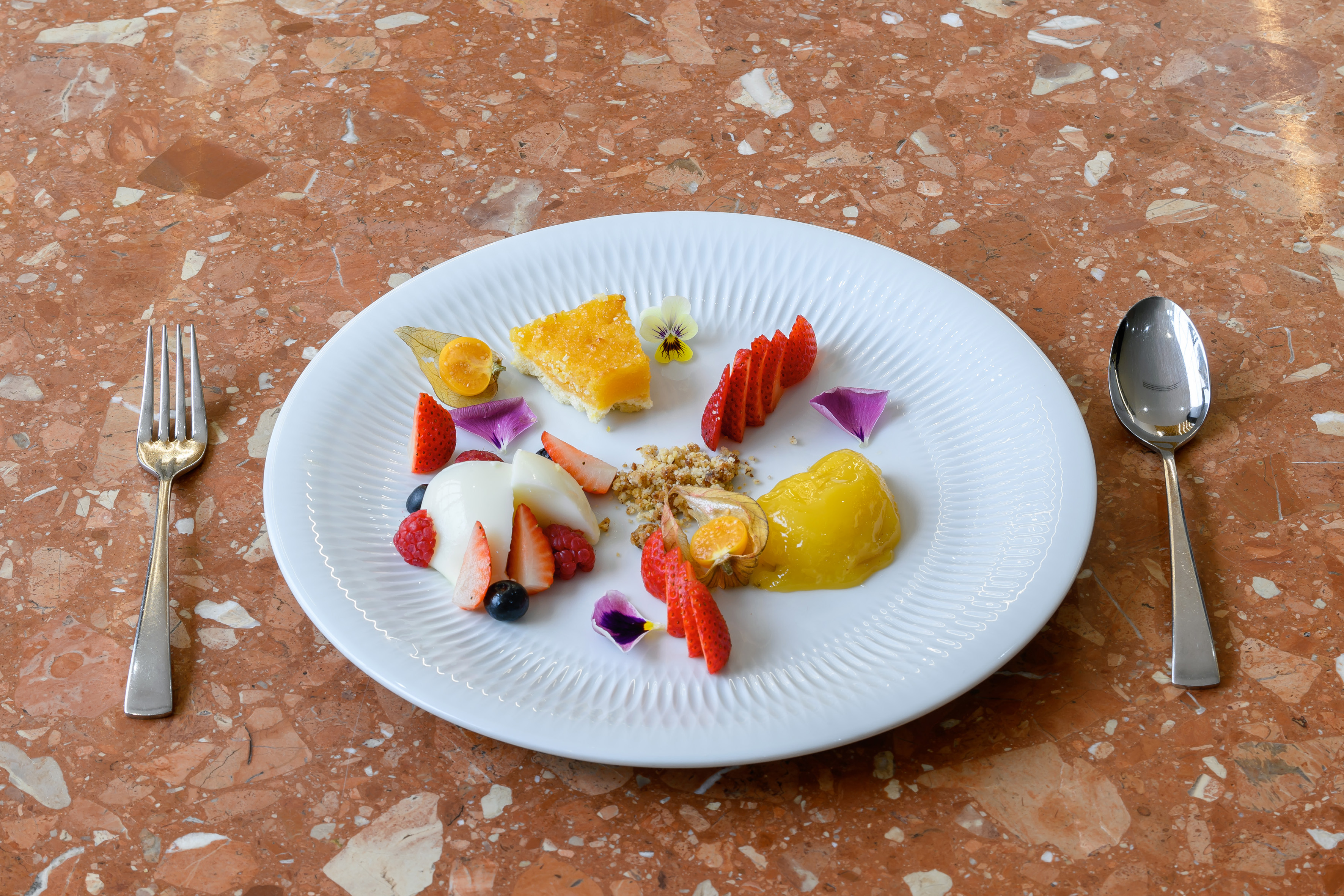 Plated dessert with cake, fresh berries, cream, and flowers on marble table.