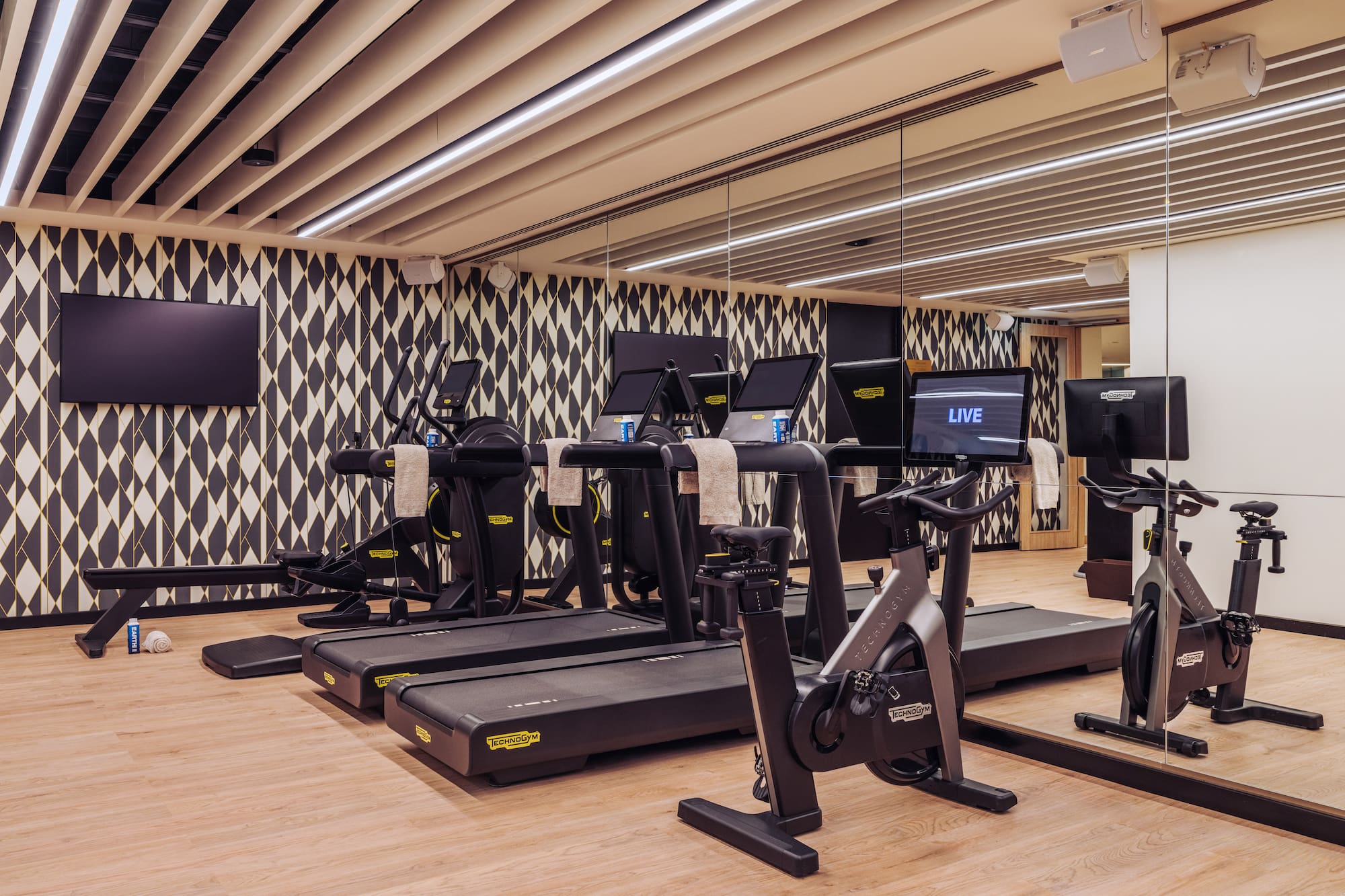 Modern gym with Technogym treadmills, bikes, rower, and geometric wall.