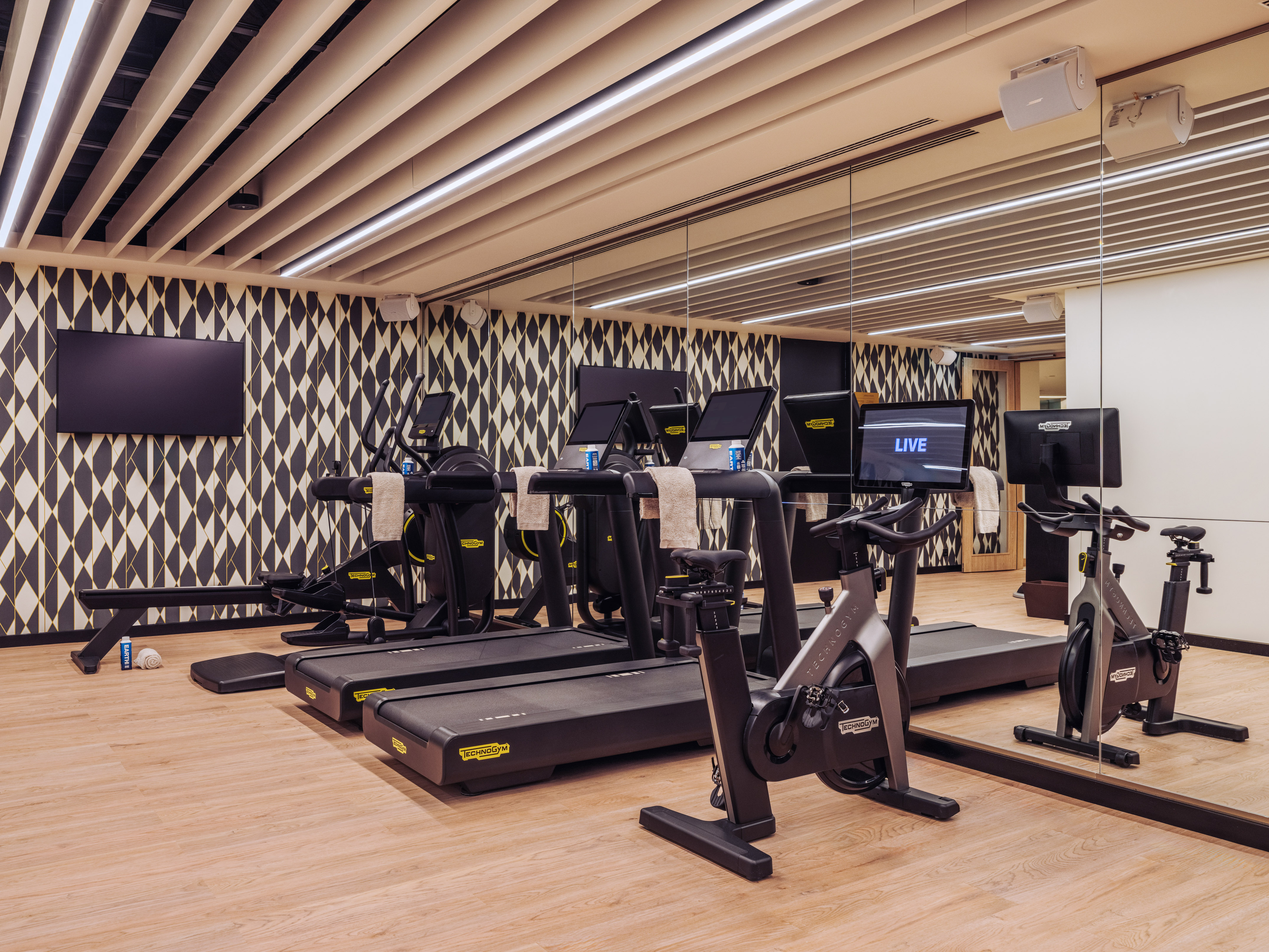 Modern gym with Technogym treadmills, bikes, rower, and geometric wall.