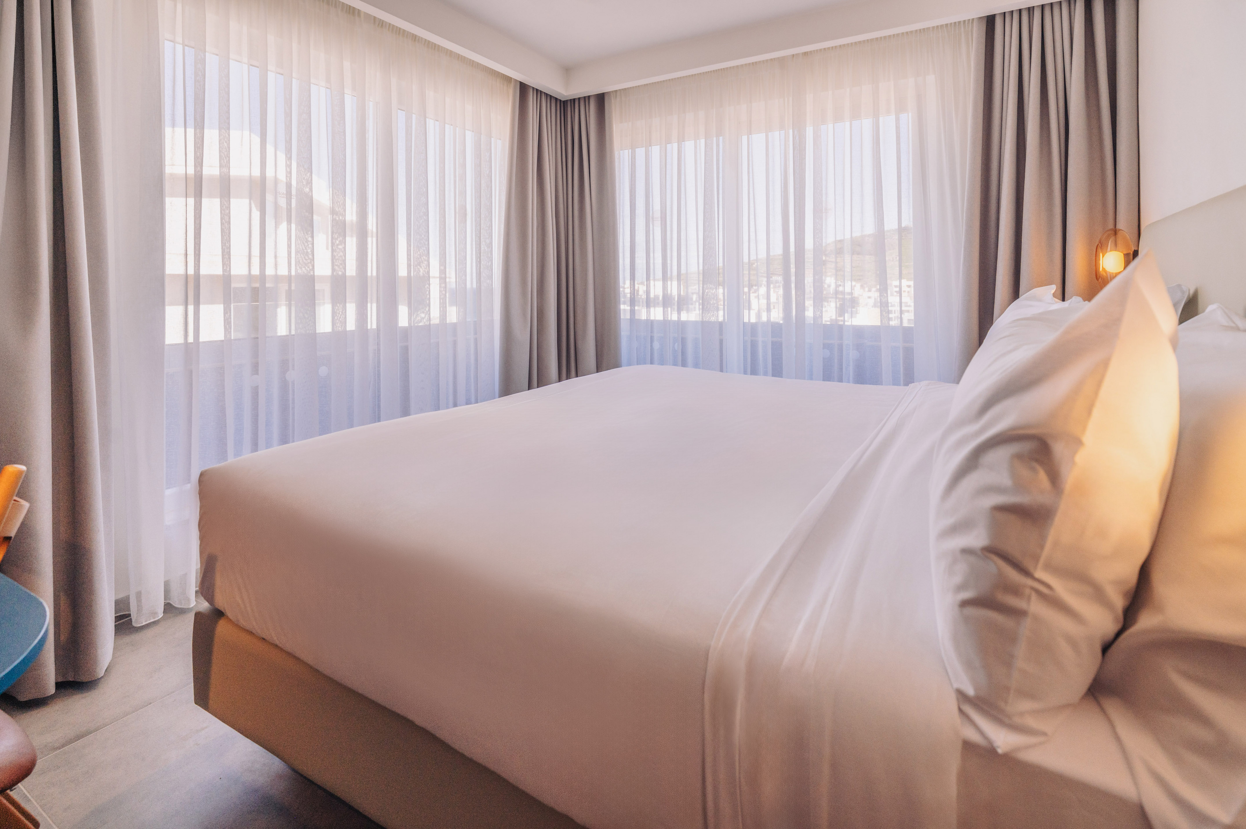 Minimalist hotel bedroom with white bed, large window, and sheer curtains.
