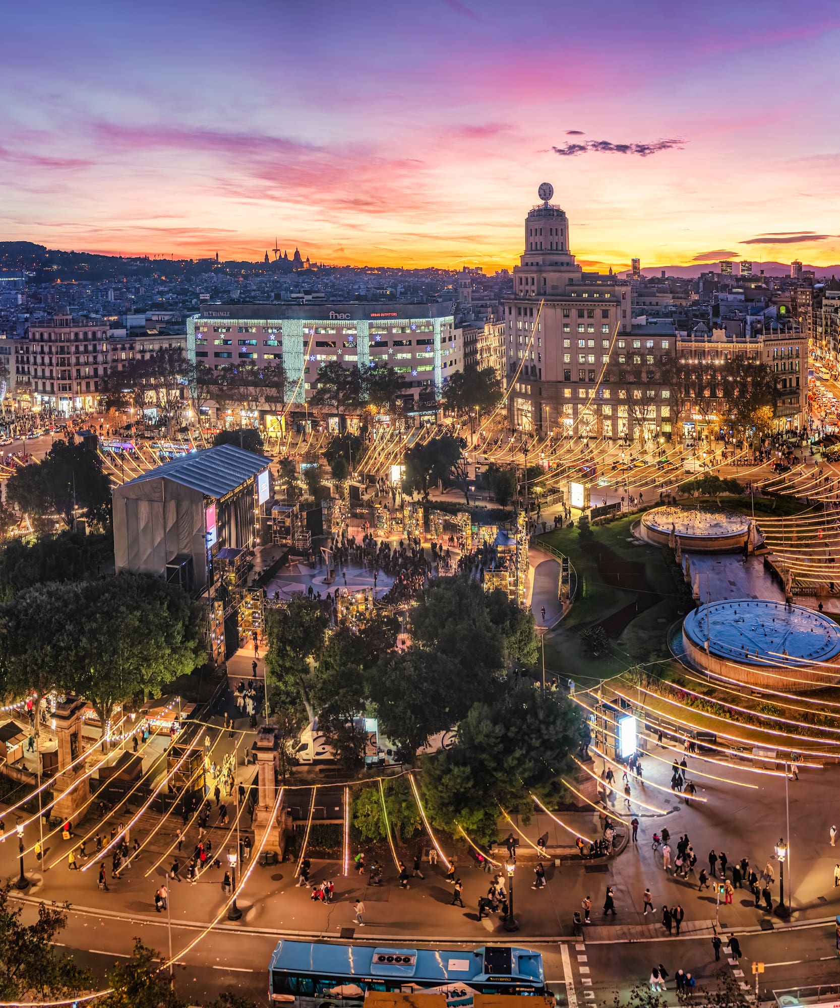 Barcelona Plaça de Catalunya at sunset, illuminated with festive lights and crowds.