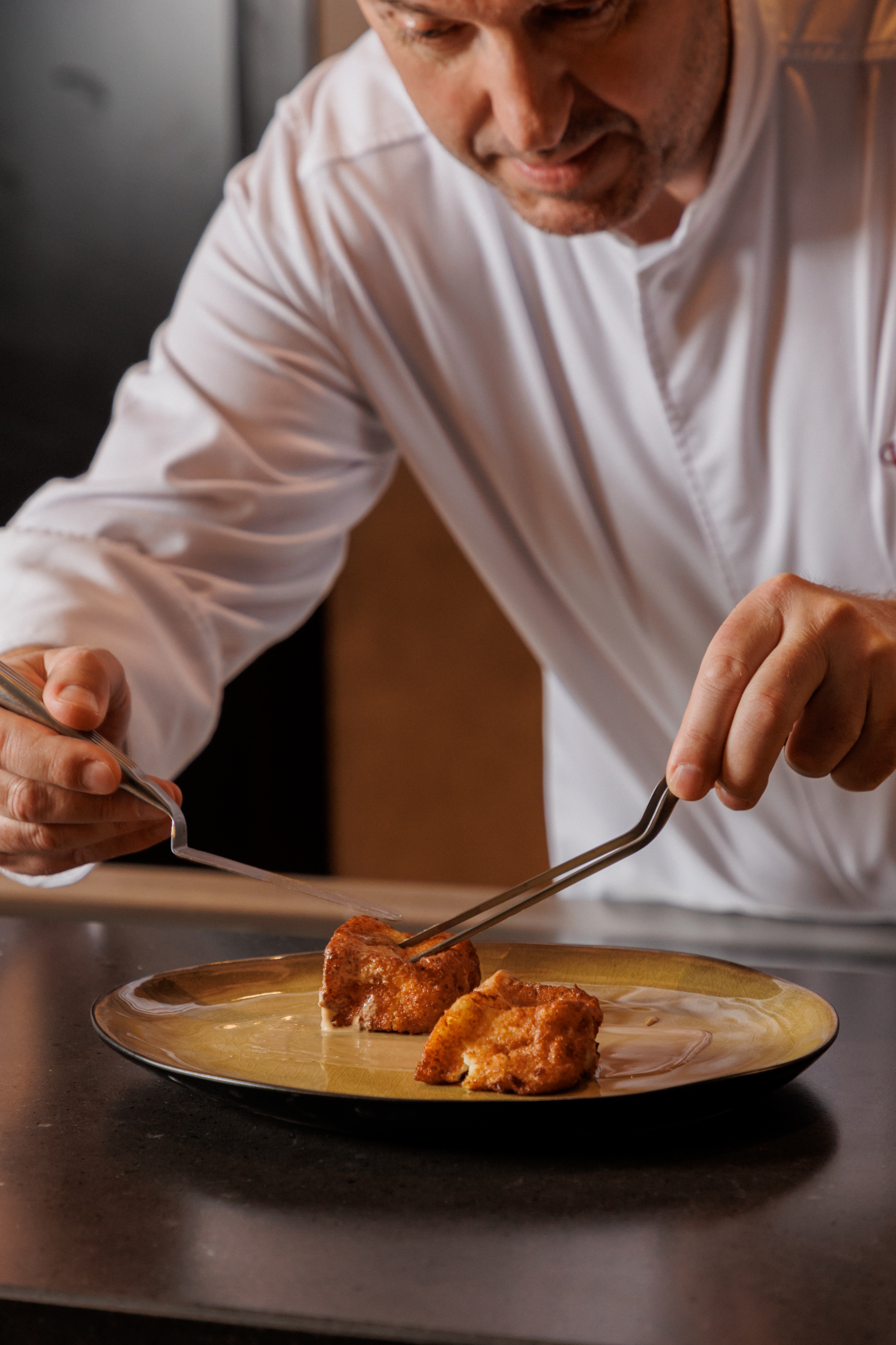 Chef in white coat plating golden fried dessert with precision tongs.