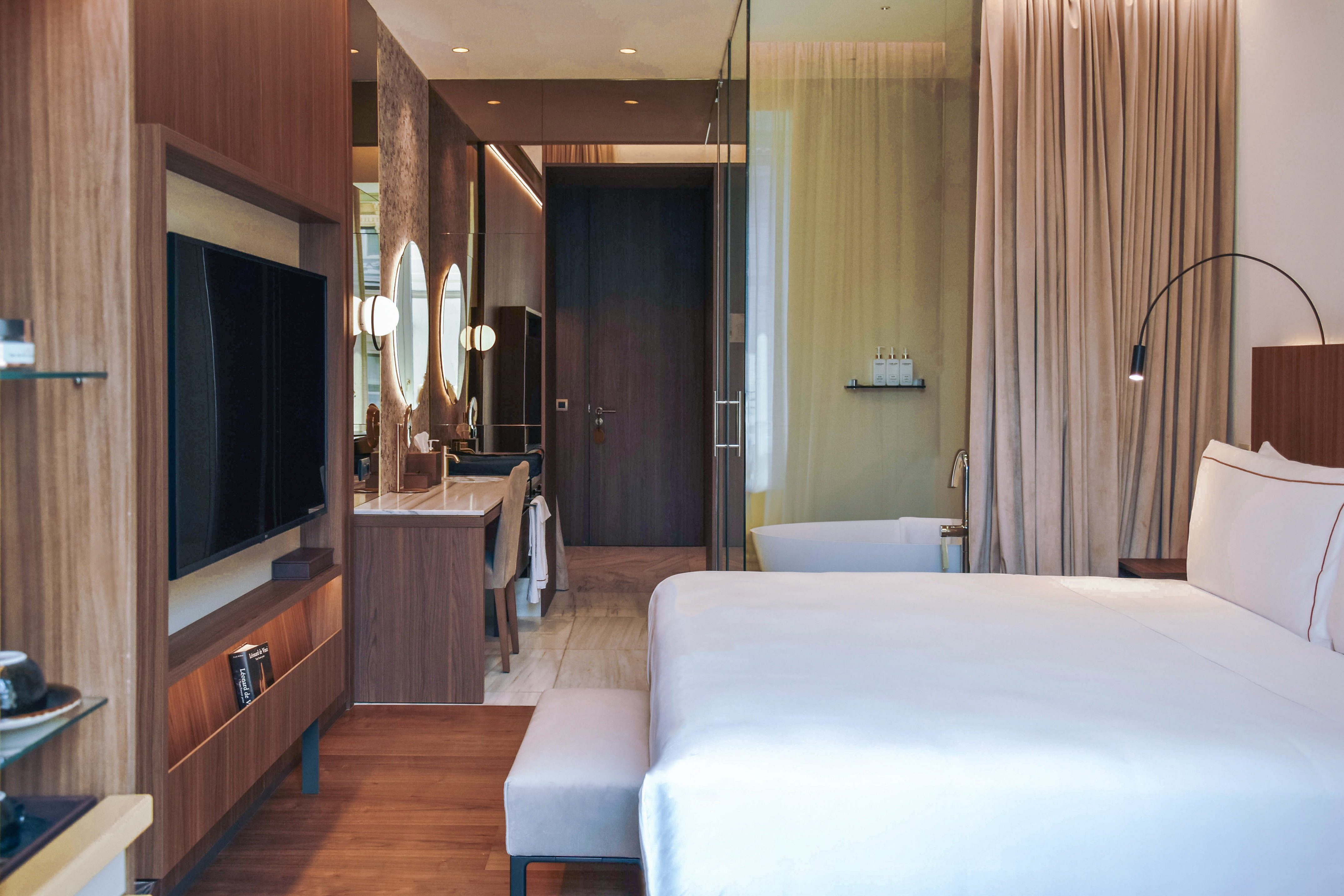 Modern hotel suite with bed, TV, vanity, and open-concept bathroom with tub.