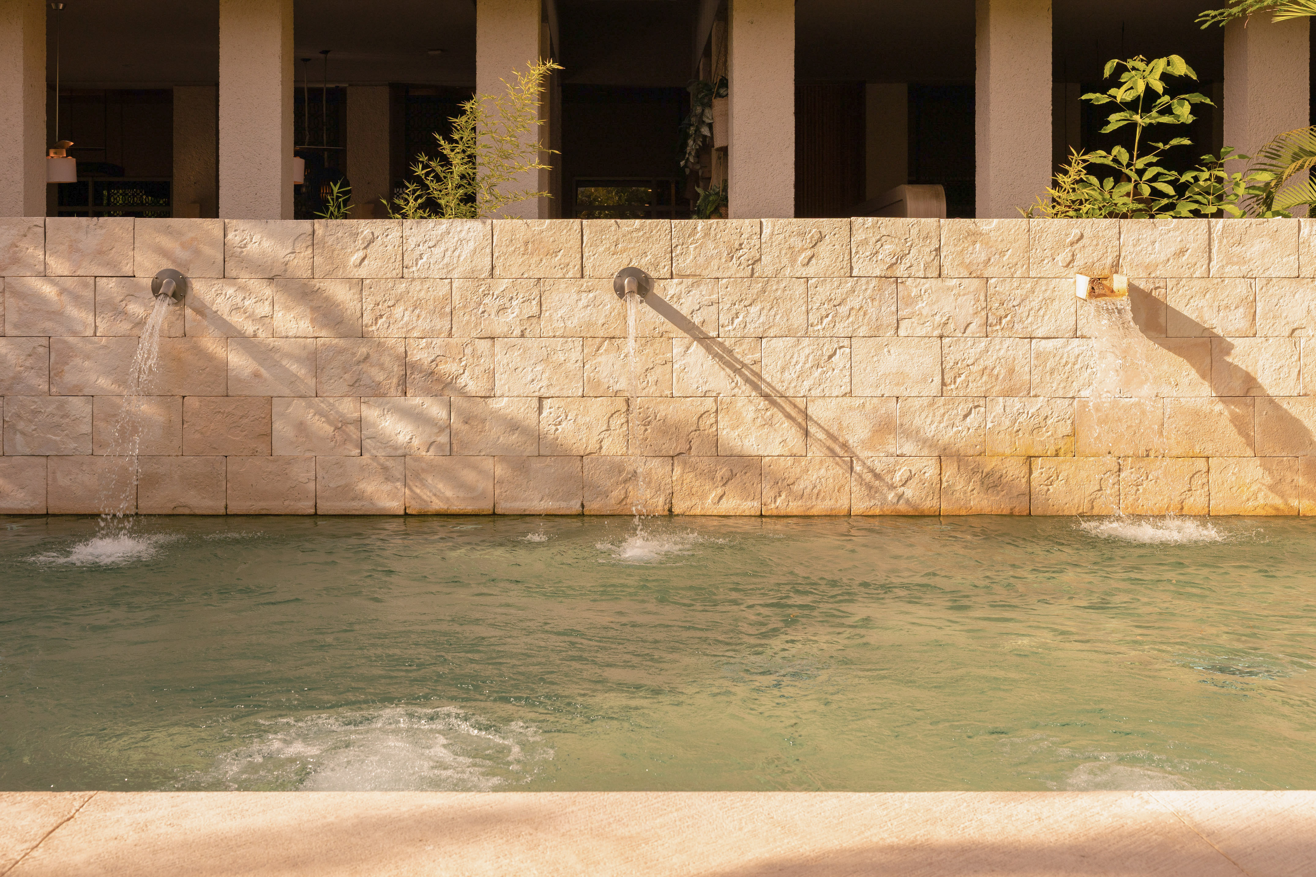 Stone wall water feature with spouts flowing into a pool, modern building background.