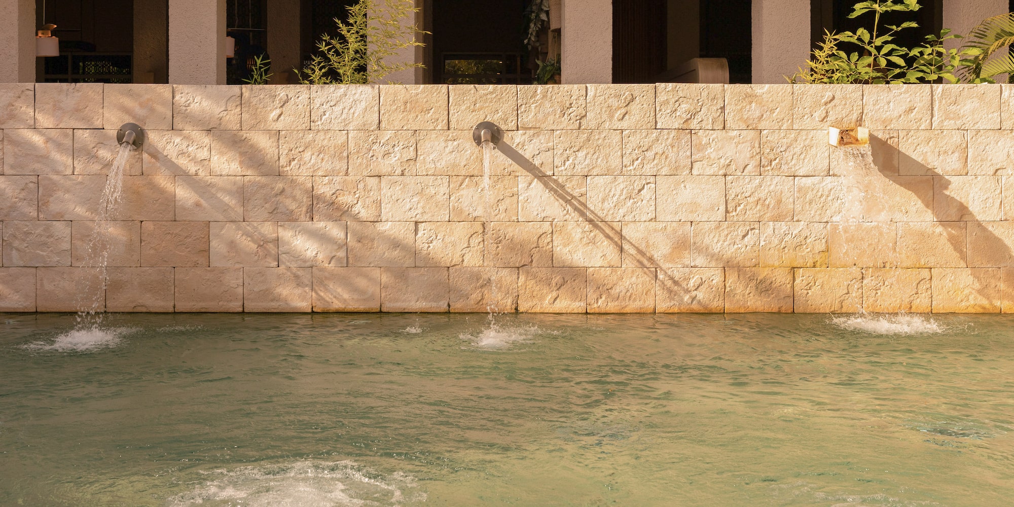 Stone wall water feature with spouts flowing into a pool, modern building background.