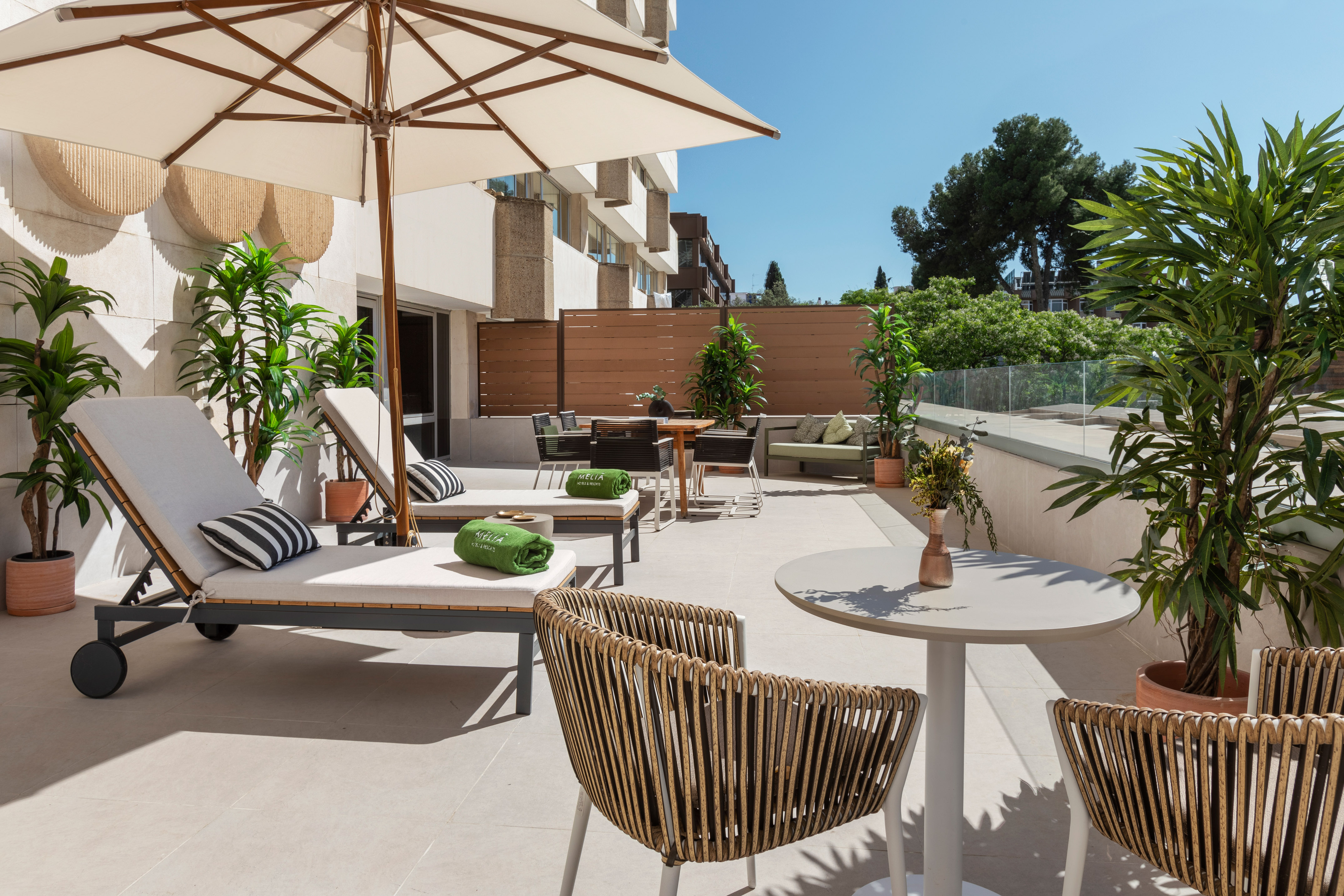 Sunny resort patio with sun loungers, umbrella, outdoor dining, and green plants.