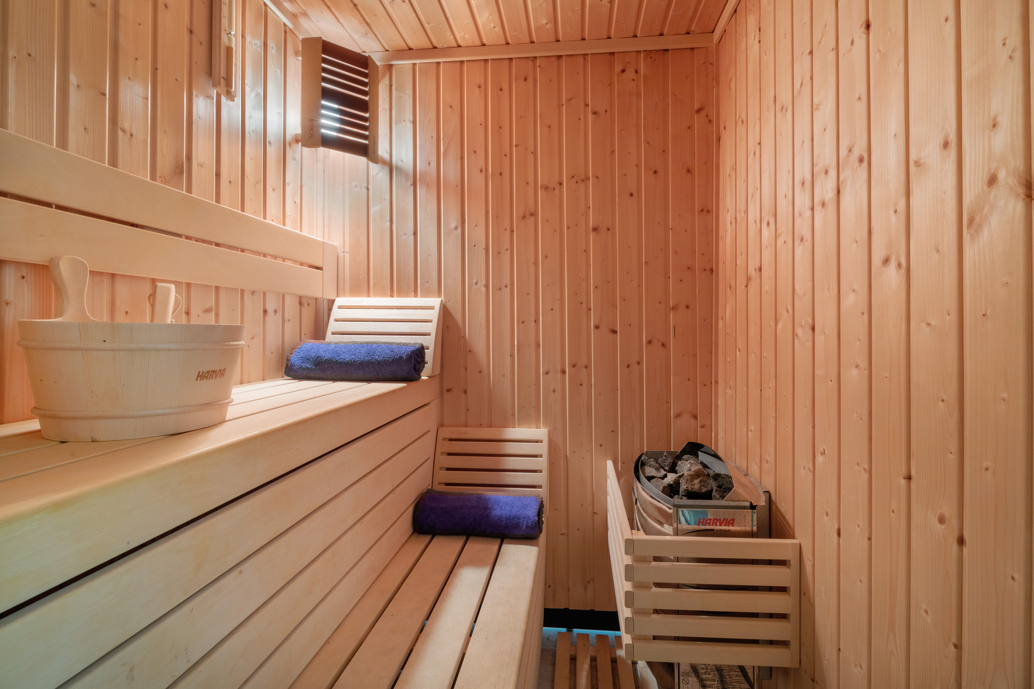 Wooden sauna interior with Harvia heater, benches, bucket, and blue towels.