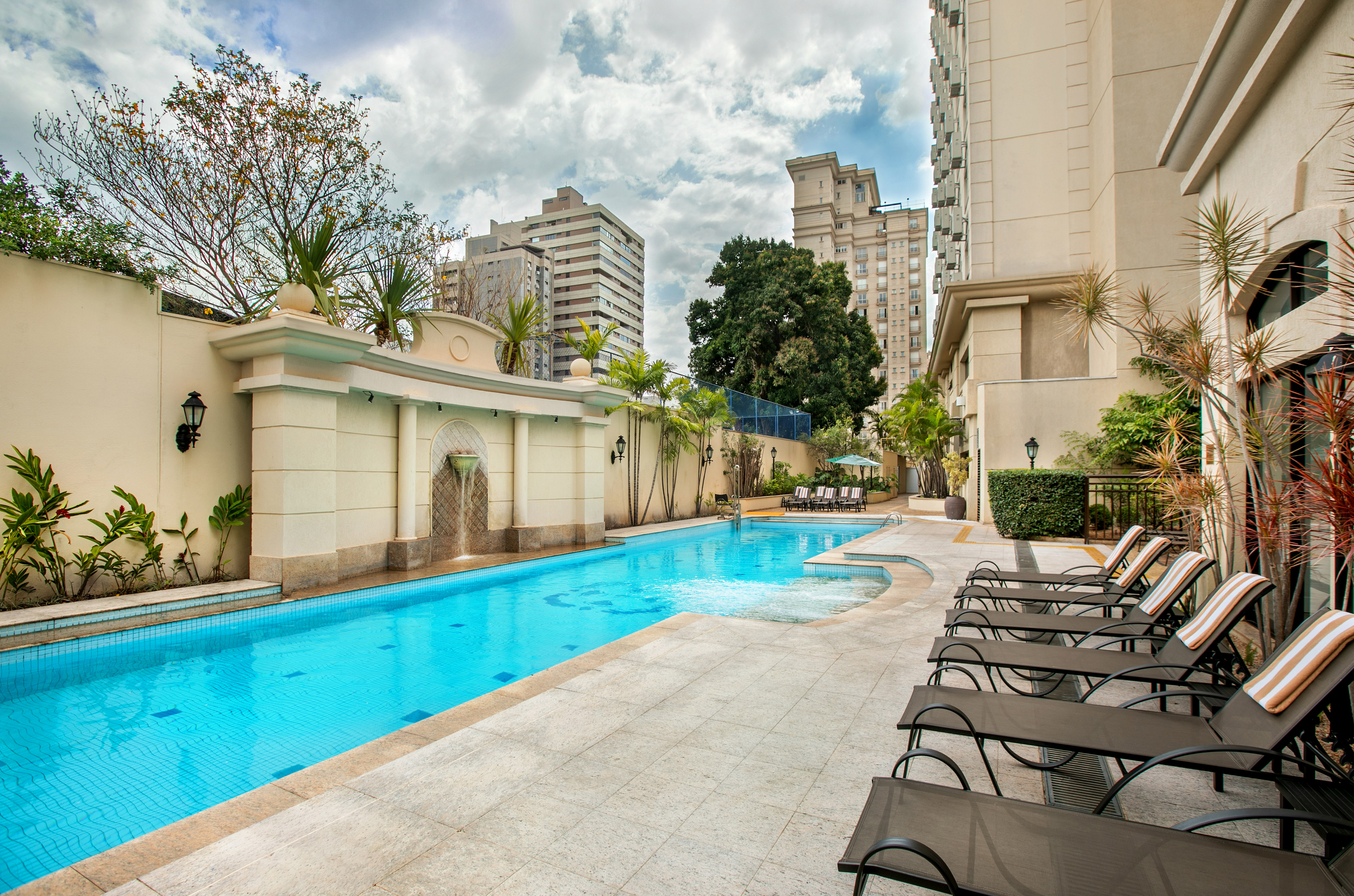 Outdoor pool with waterfall, lounge chairs, and modern buildings under a cloudy sky.