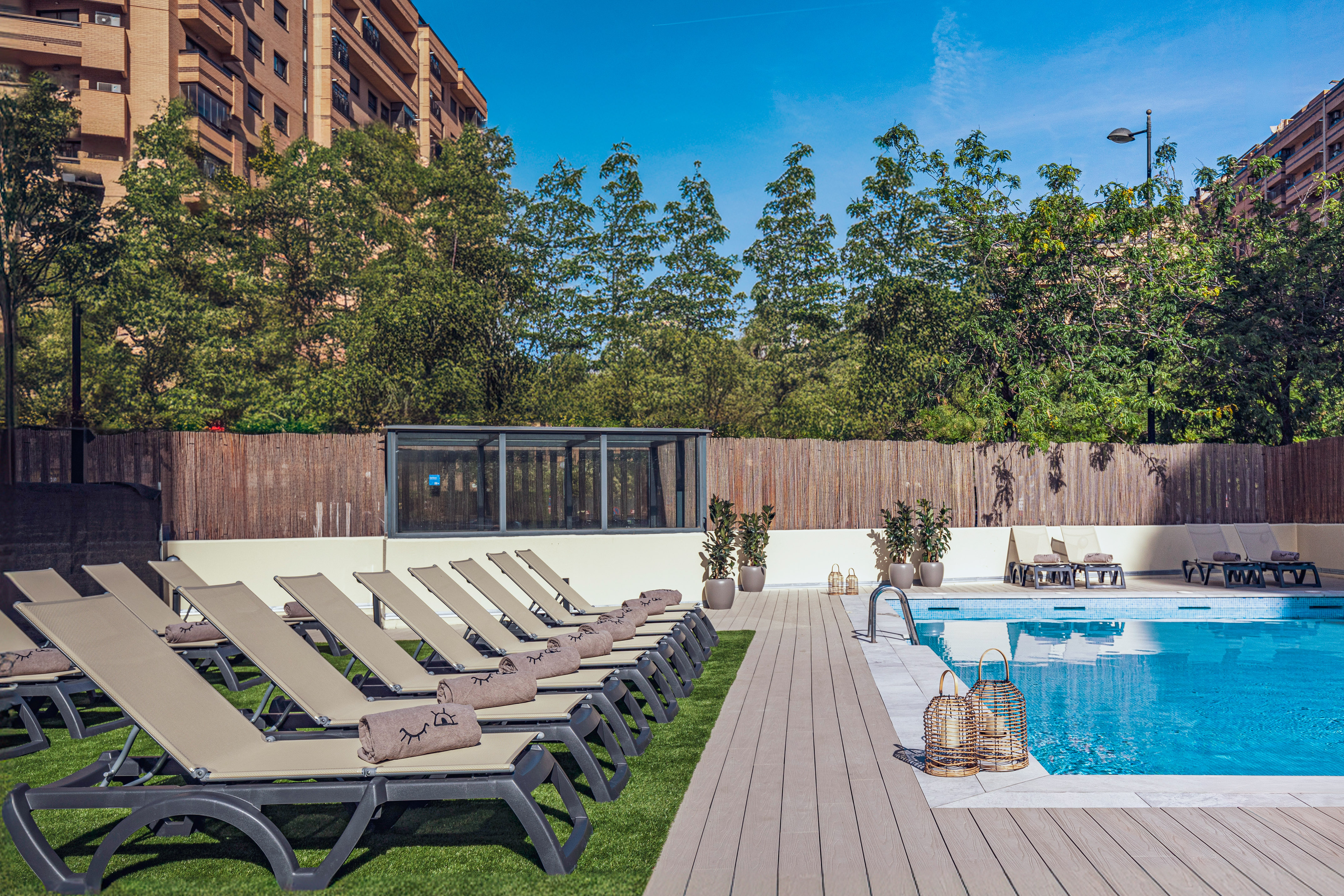 Sunny hotel swimming pool area with lounge chairs and green trees.