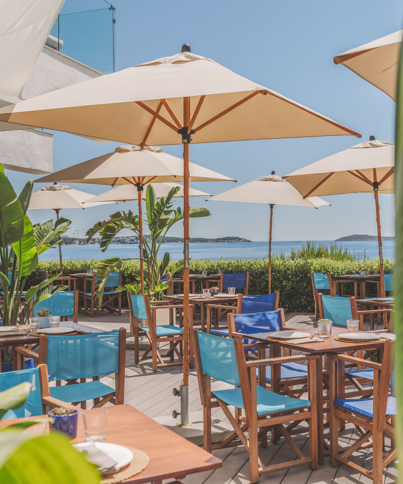 Seaside restaurant patio with blue chairs, beige umbrellas, and ocean.