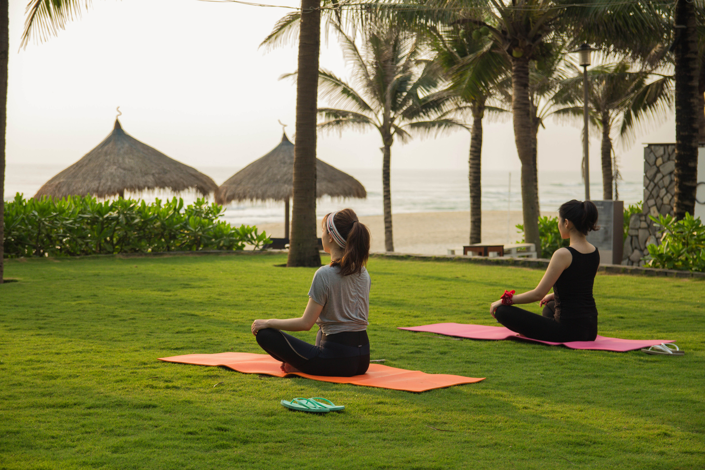 two women sitting on yoga mats on grass with palm trees and straw umbrellas