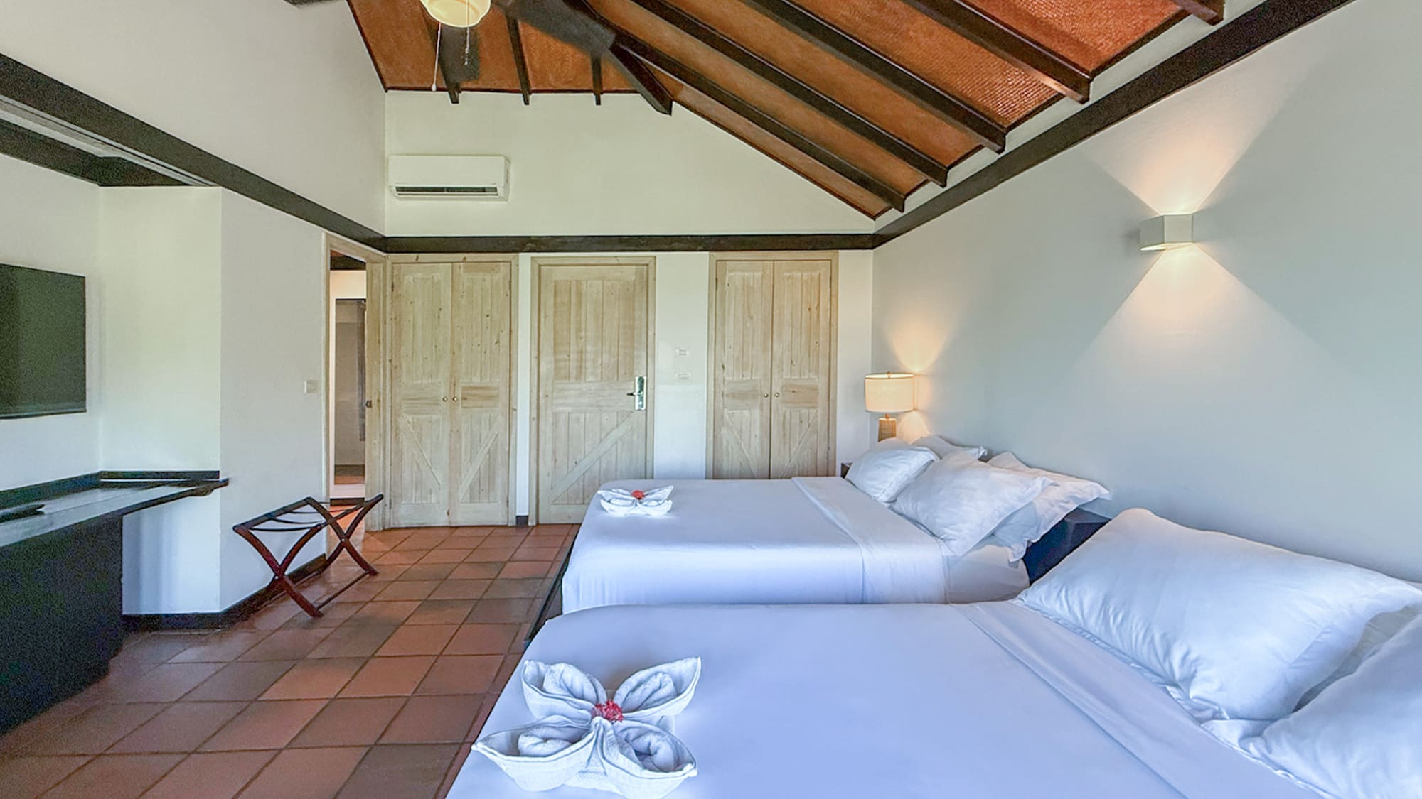 Resort bedroom with two white beds, vaulted wooden ceiling, and terracotta tiled floor.