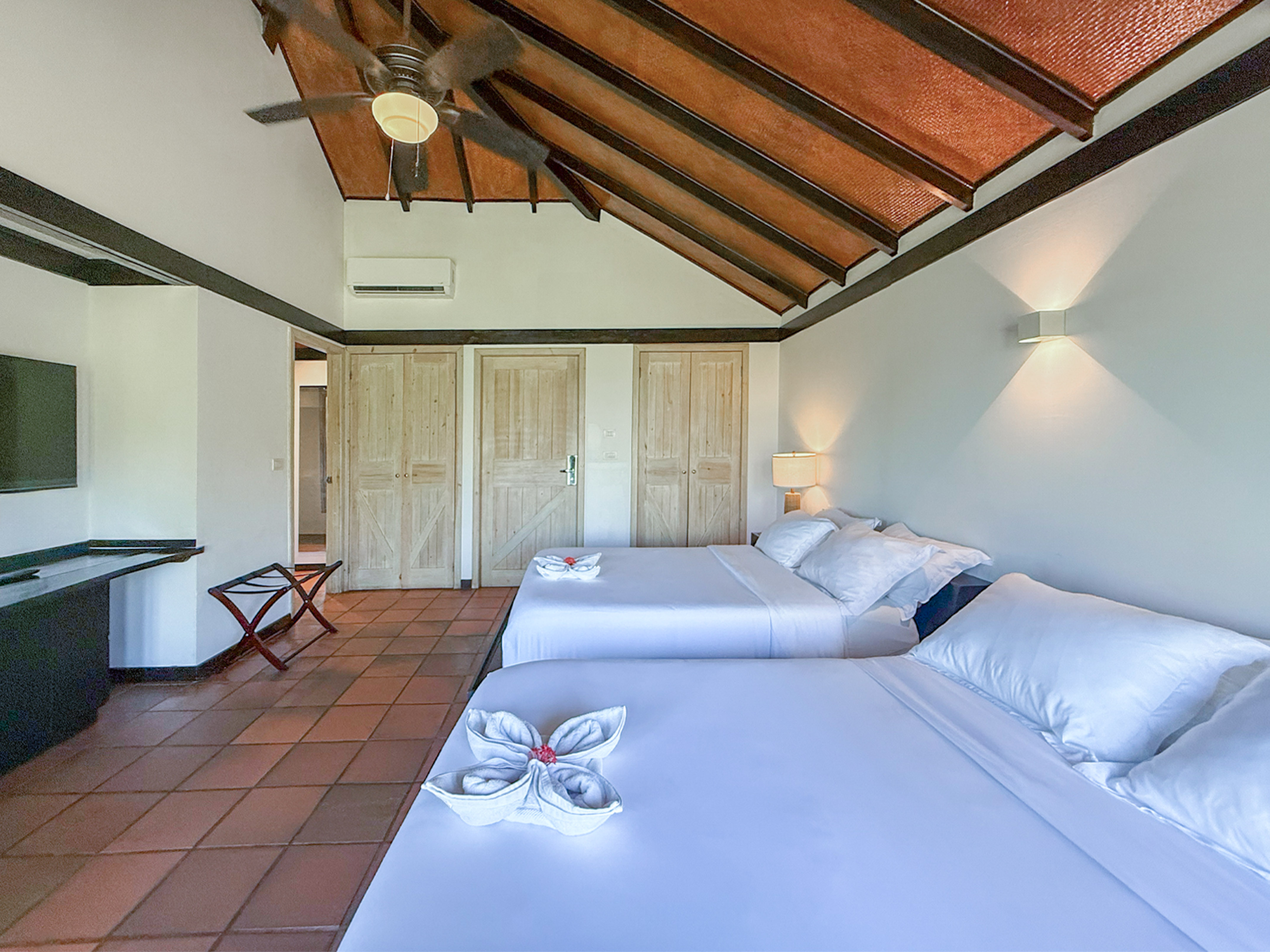 Resort bedroom with two white beds, vaulted wooden ceiling, and terracotta tiled floor.
