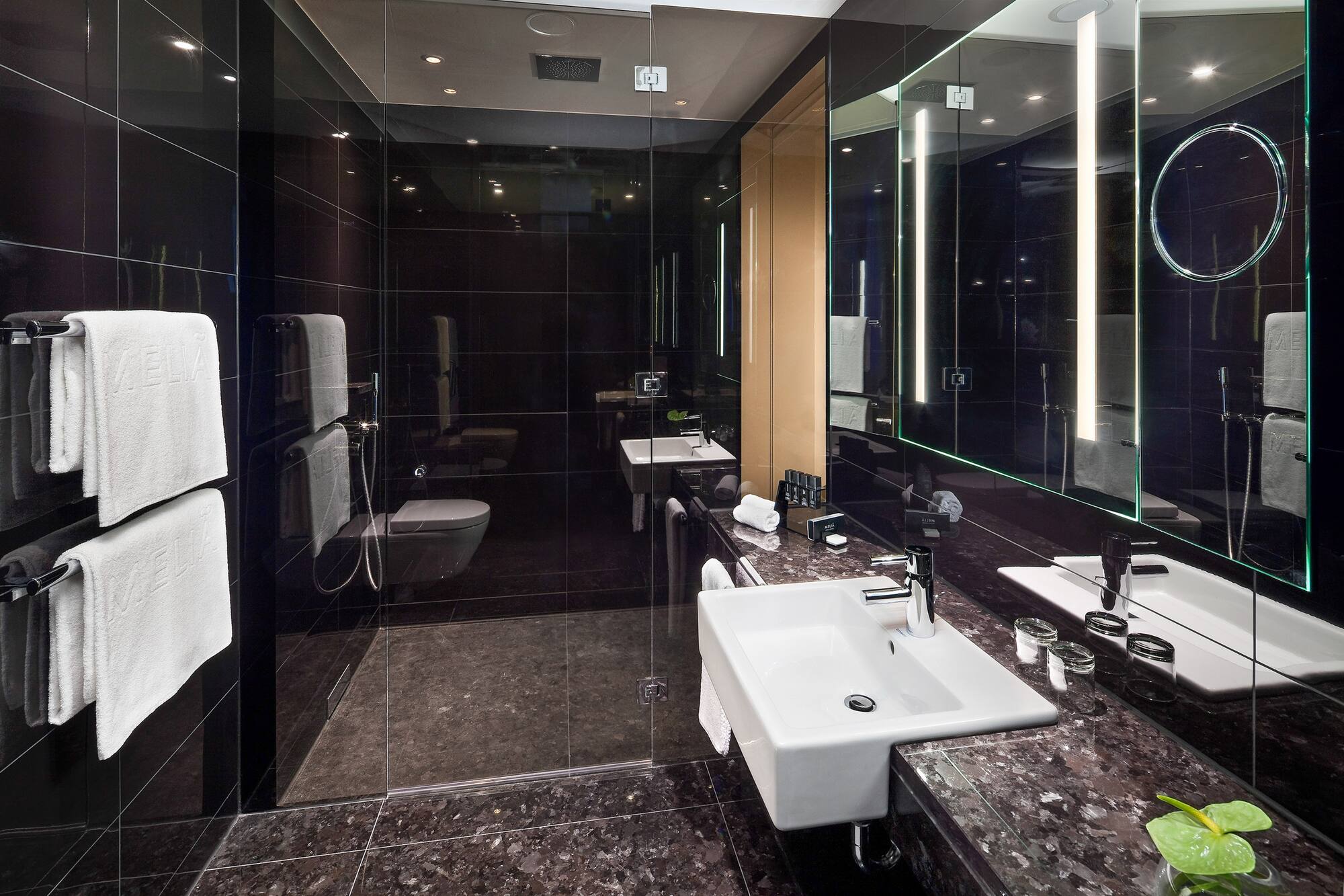 Black tiled hotel bathroom with glass shower, white sink, and backlit mirror.