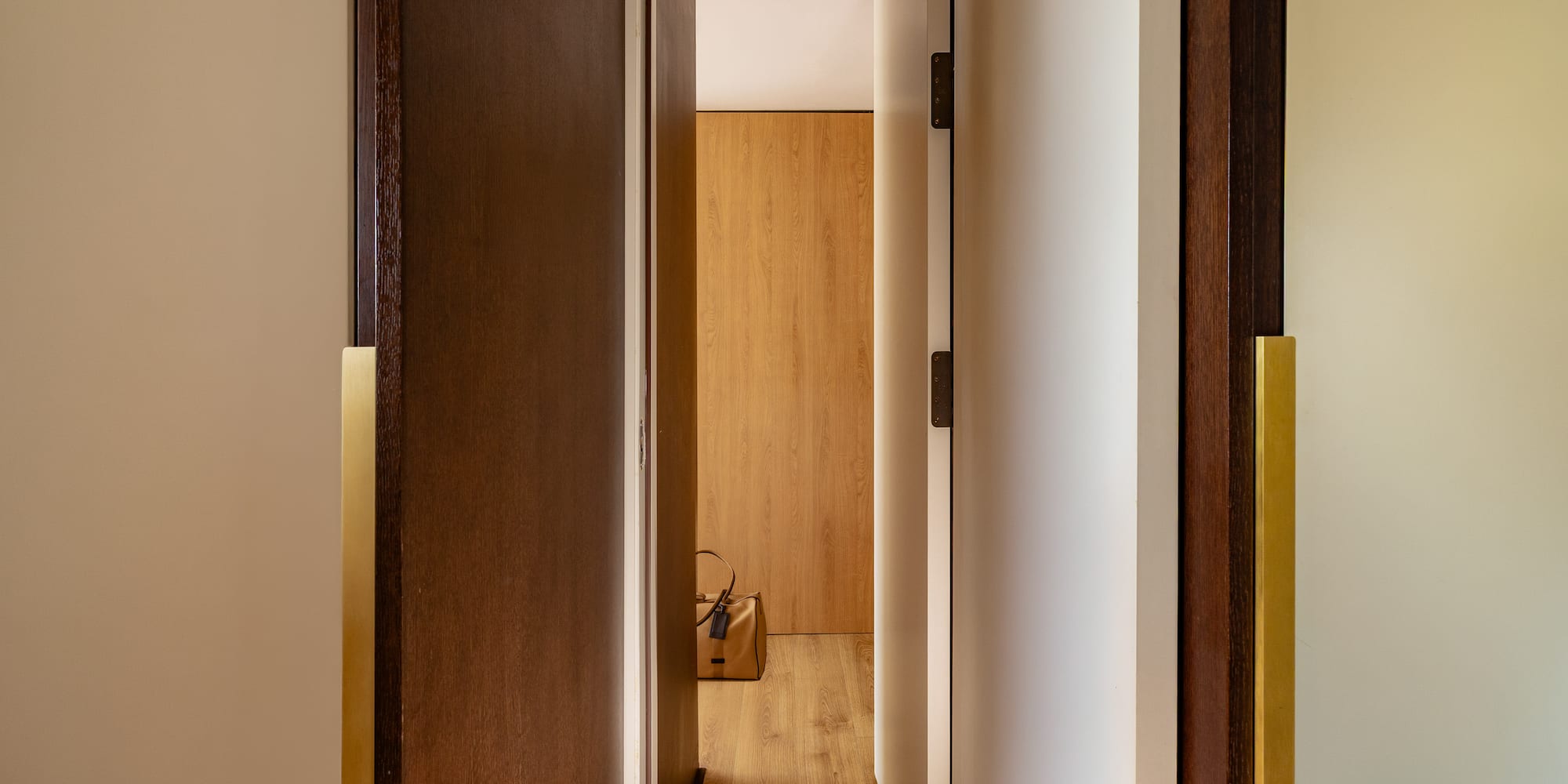 Modern hallway with open wooden doors, brass handles, and a brown bag on light wood floor.