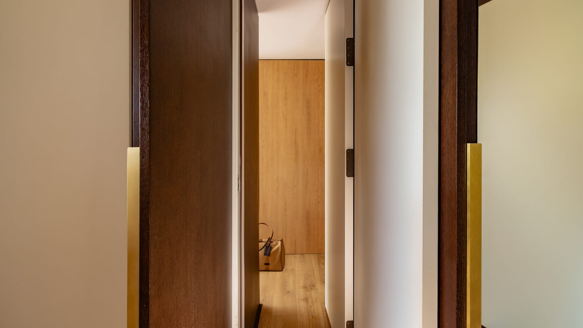 Modern hallway with open wooden doors, brass handles, and a brown bag on light wood floor.