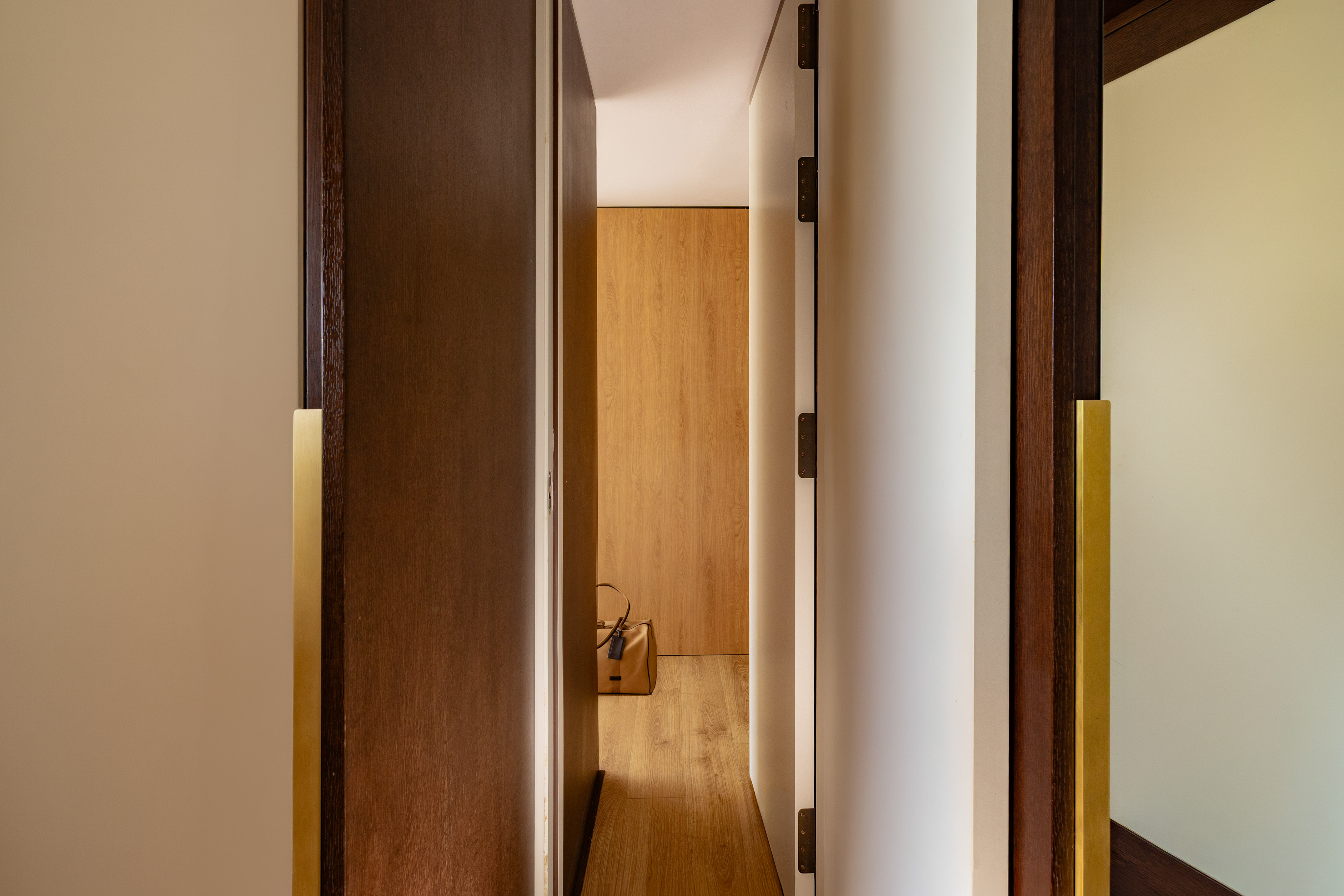 Modern hallway with open wooden doors, brass handles, and a brown bag on light wood floor.