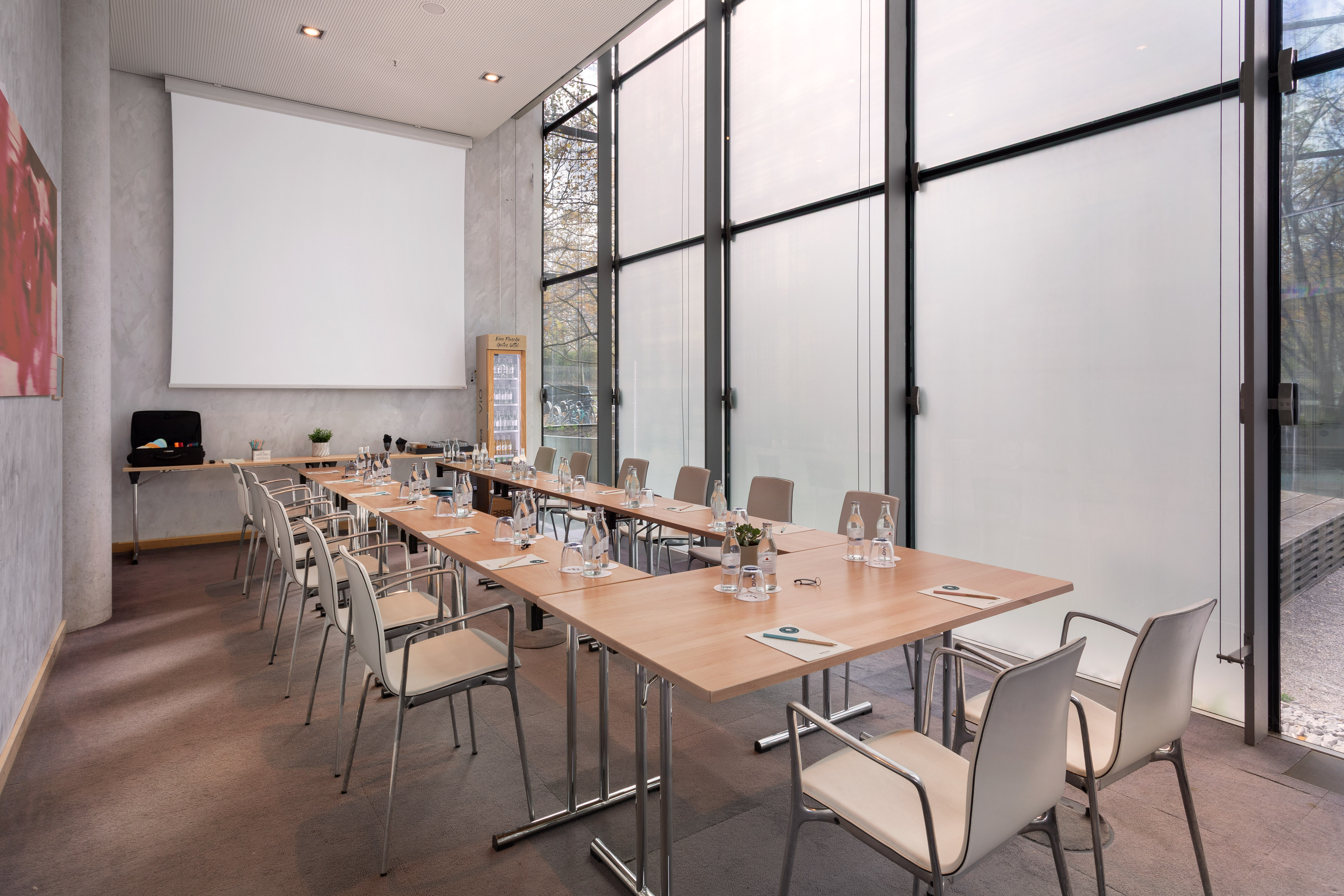 Bright conference room with U-shaped tables, screen, and large windows.