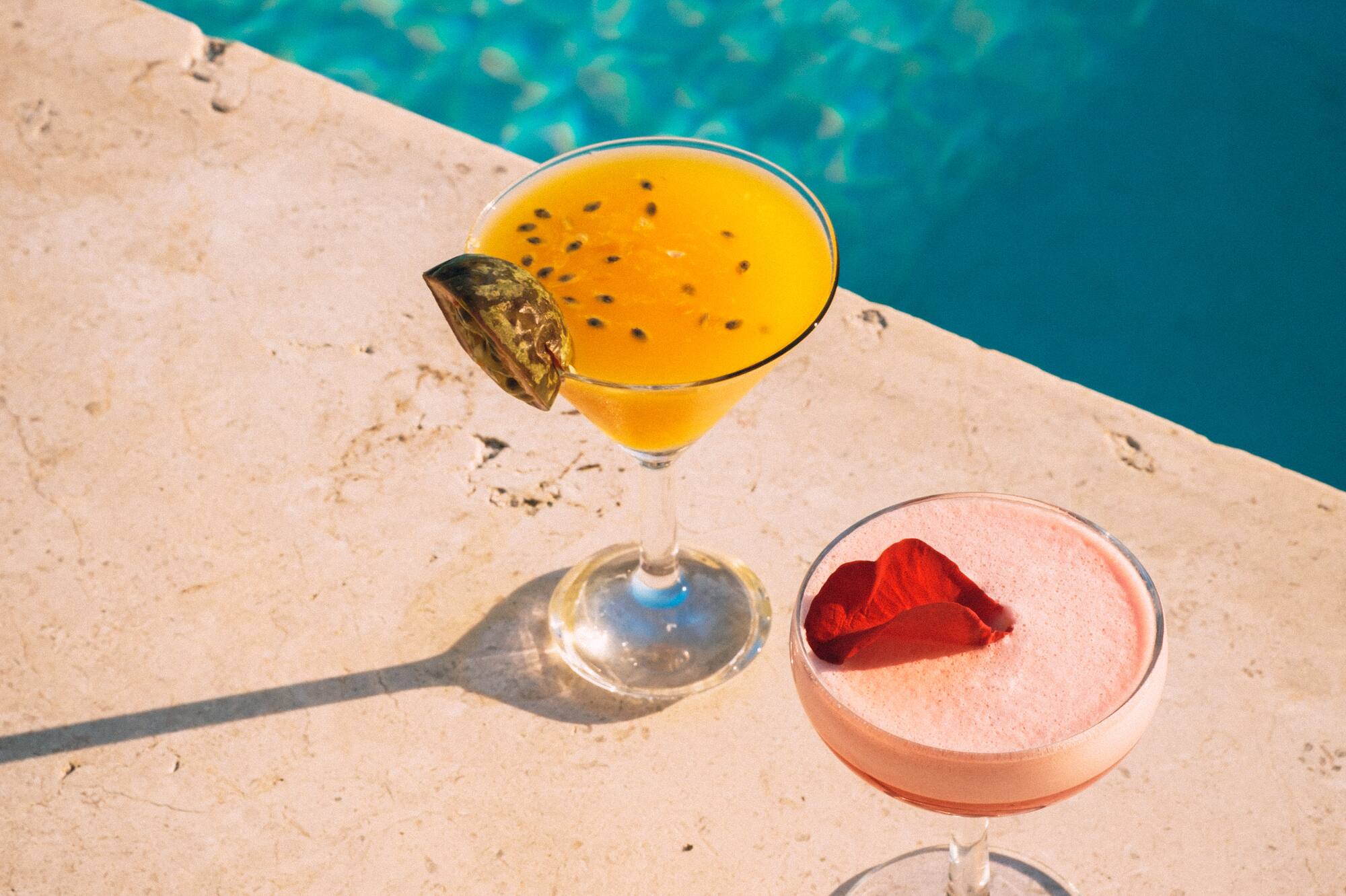 Passion fruit cocktail and pink cocktail with petal by a blue pool.