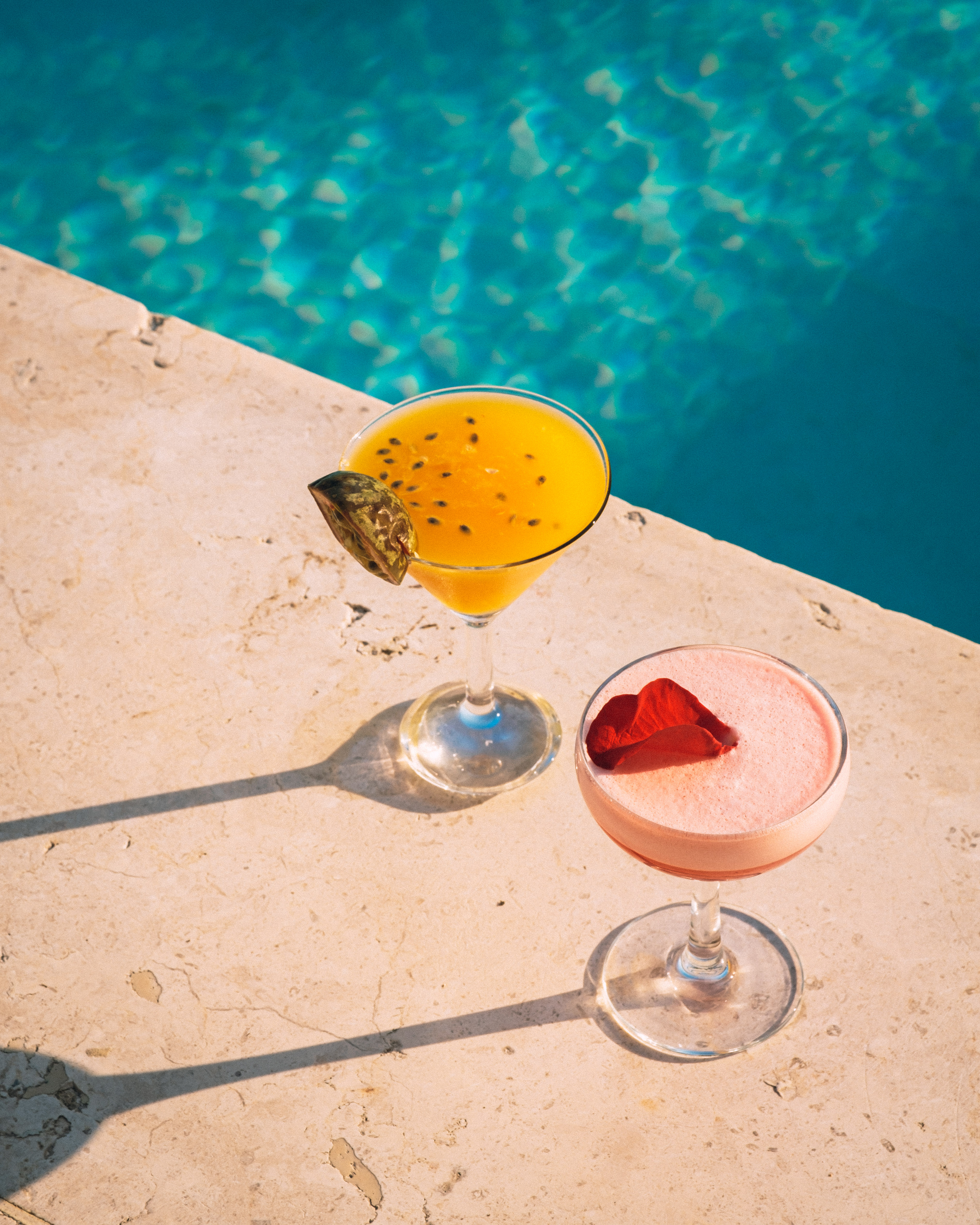 Passion fruit cocktail and pink cocktail with petal by a blue pool.