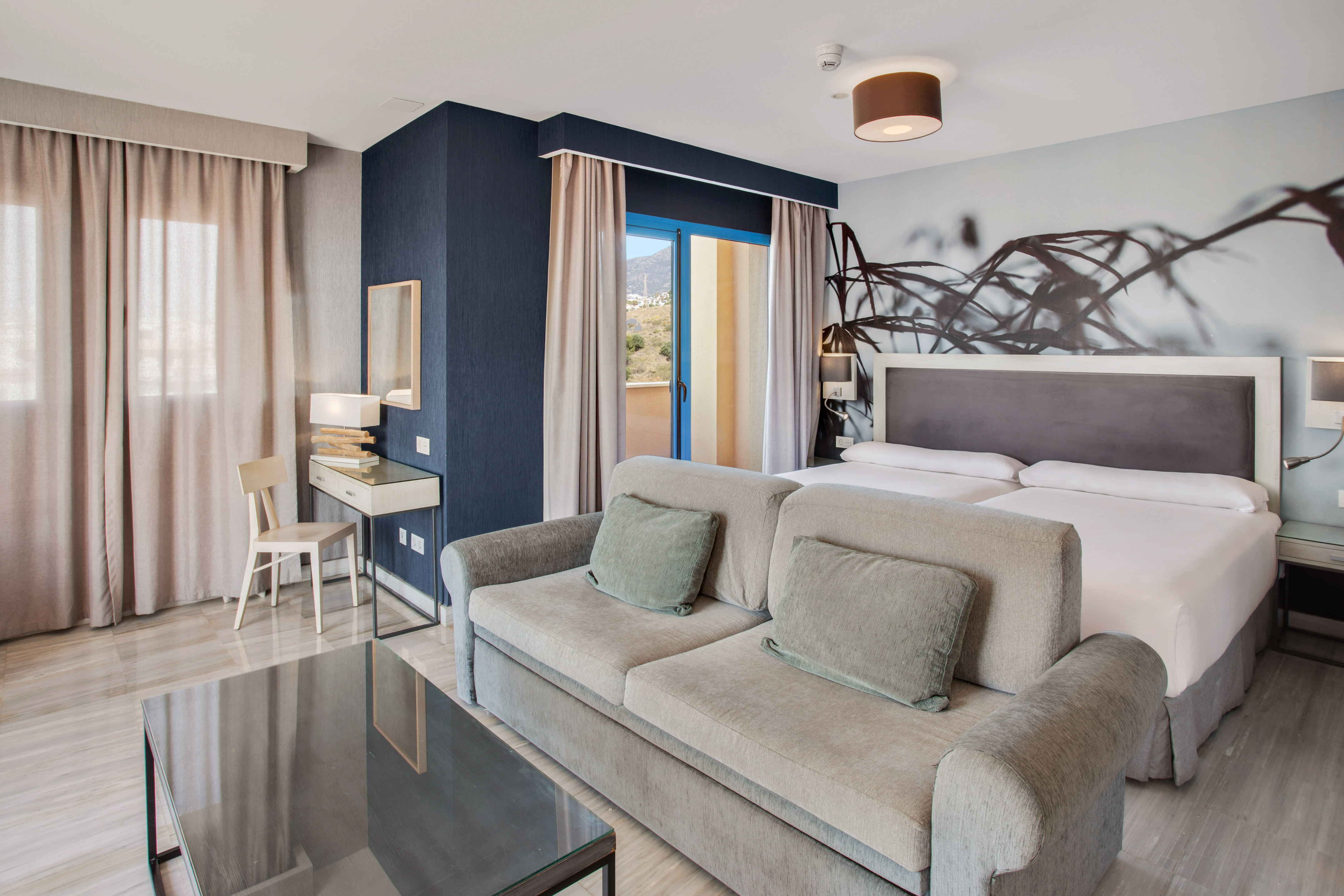 Modern hotel room with sofa, twin beds, balcony, and mountain views.