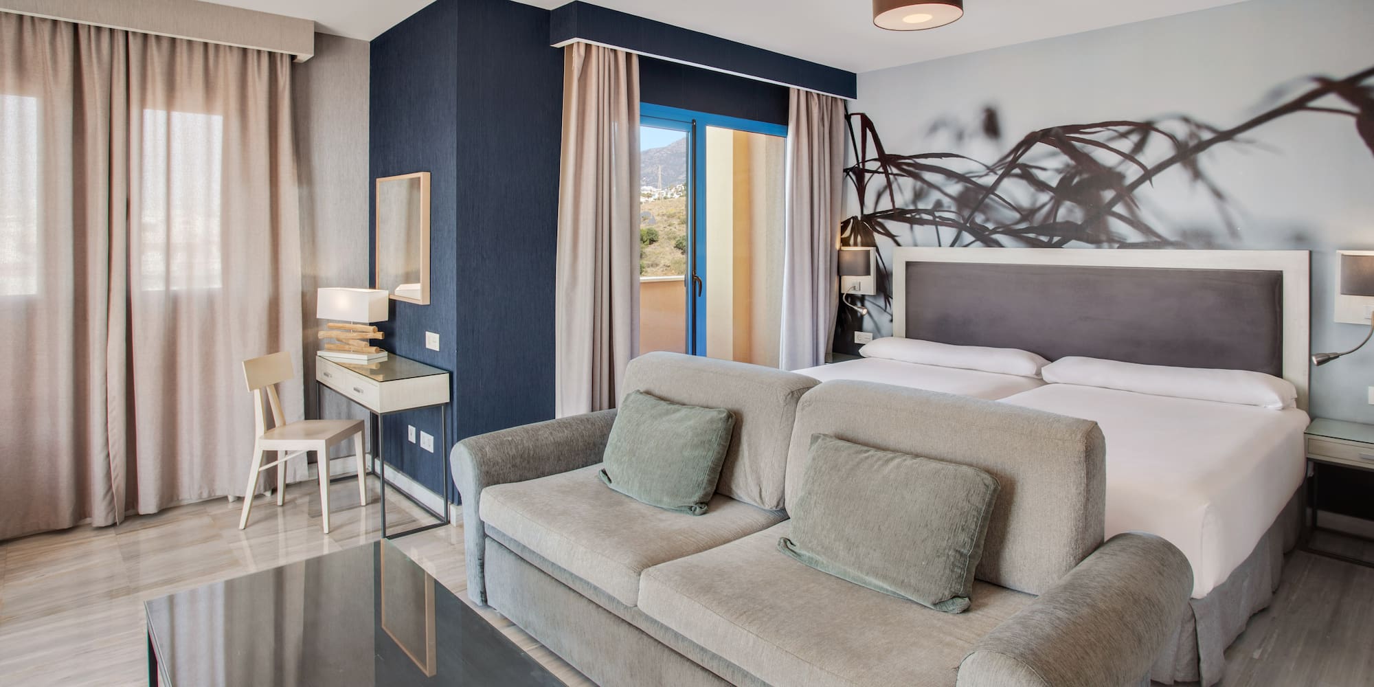 Modern hotel room with sofa, twin beds, balcony, and mountain views.