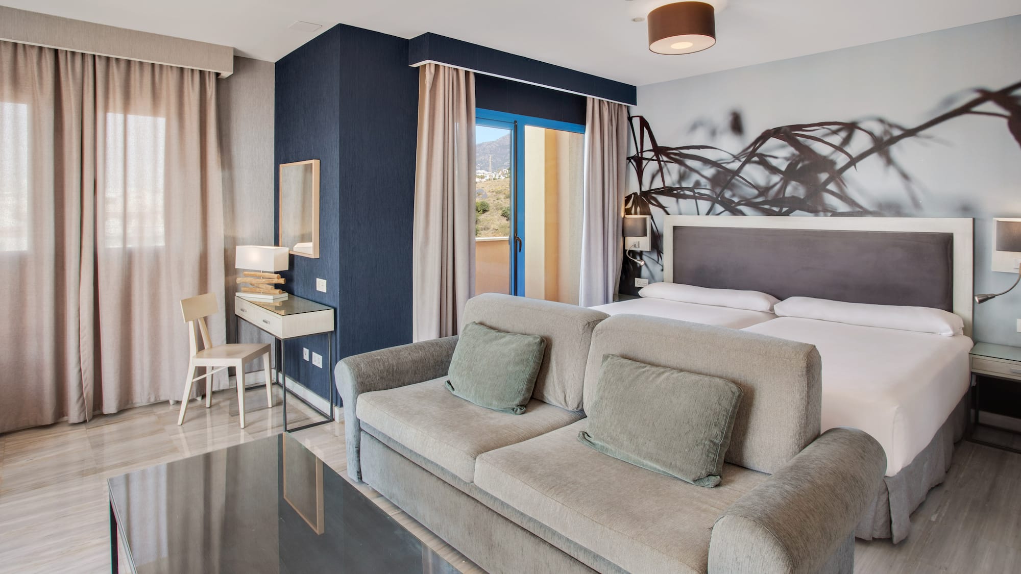 Modern hotel room with sofa, twin beds, balcony, and mountain views.