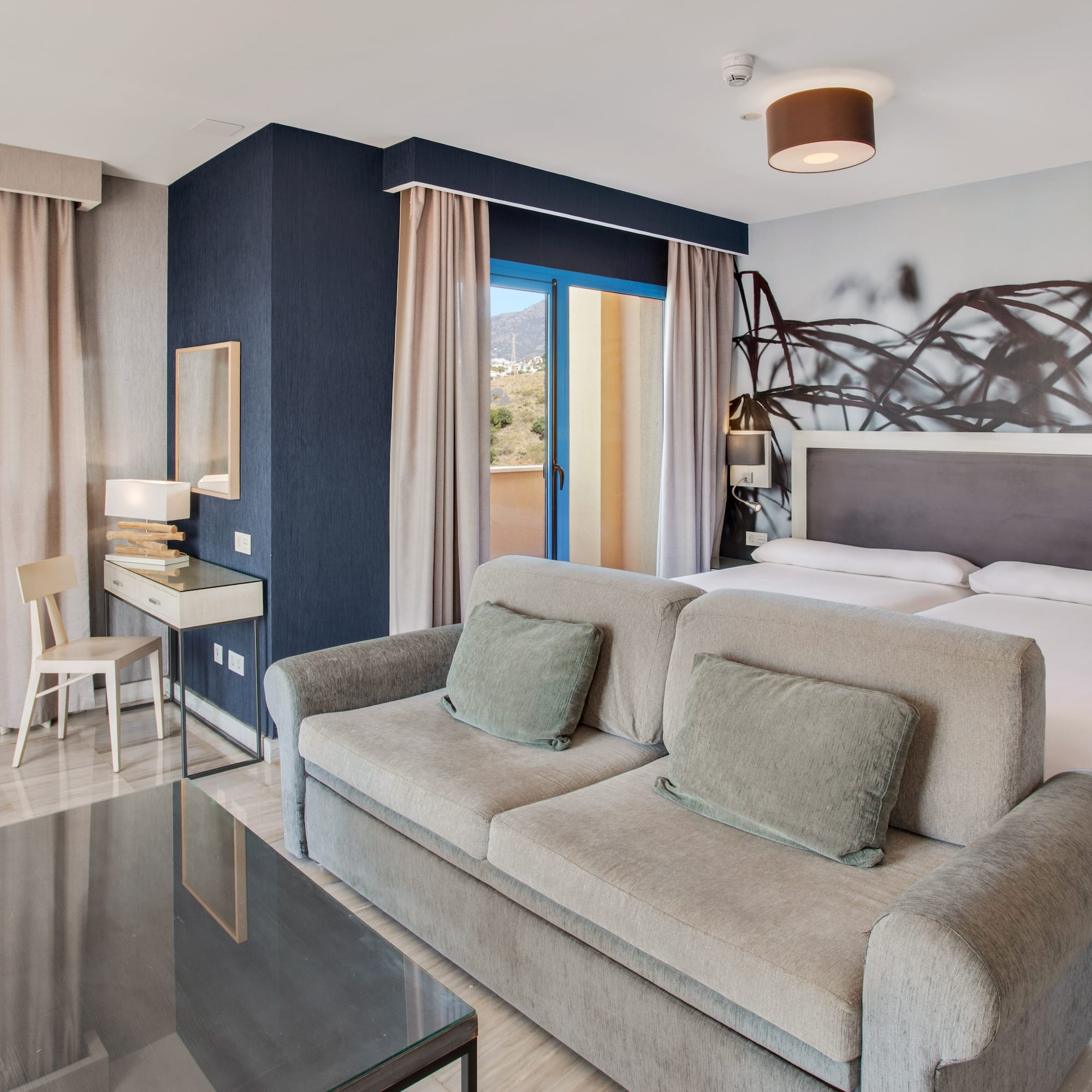 Modern hotel room with sofa, twin beds, balcony, and mountain views.