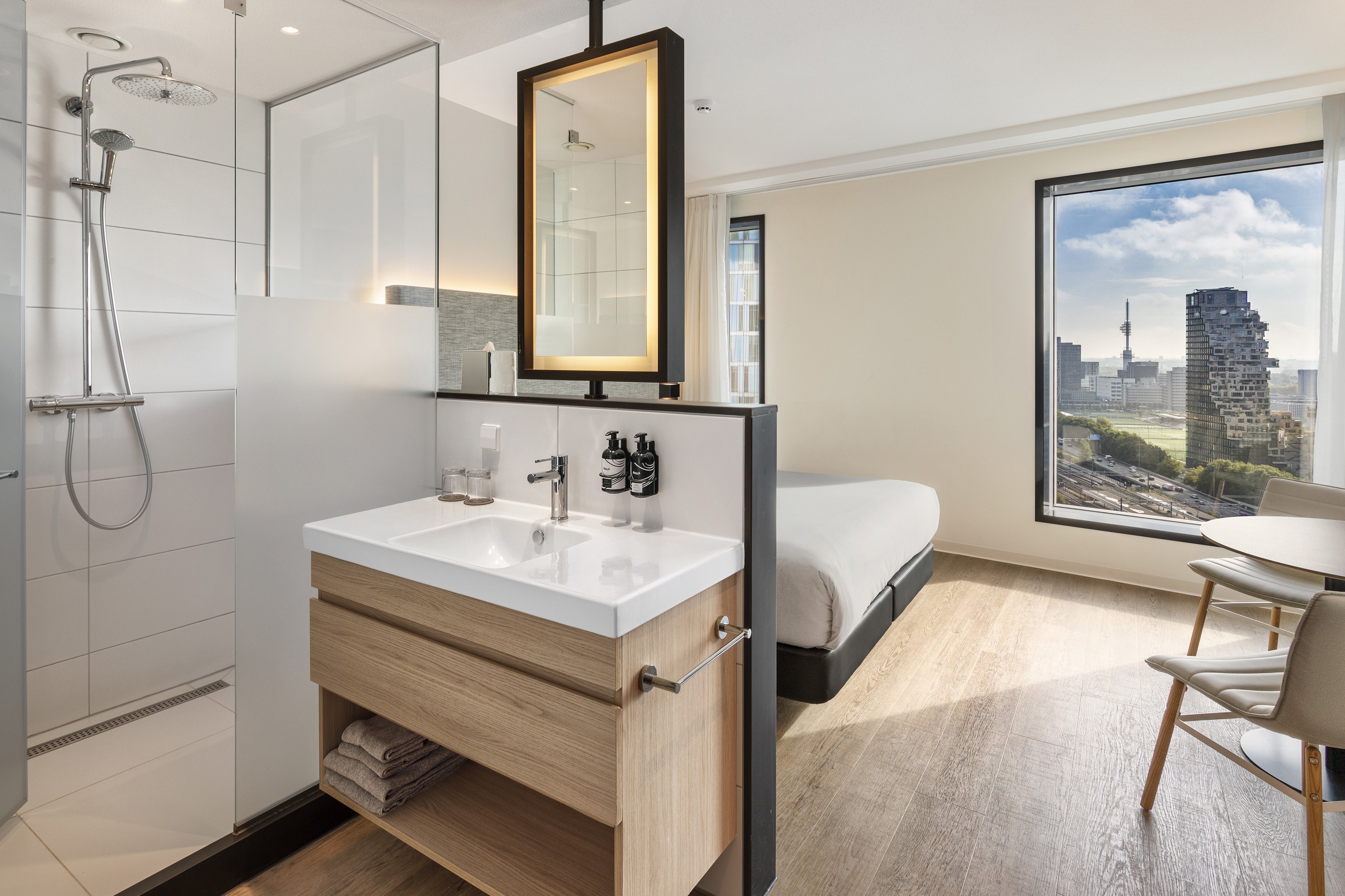 Modern hotel room with open plan shower, sink, bed, and city view.