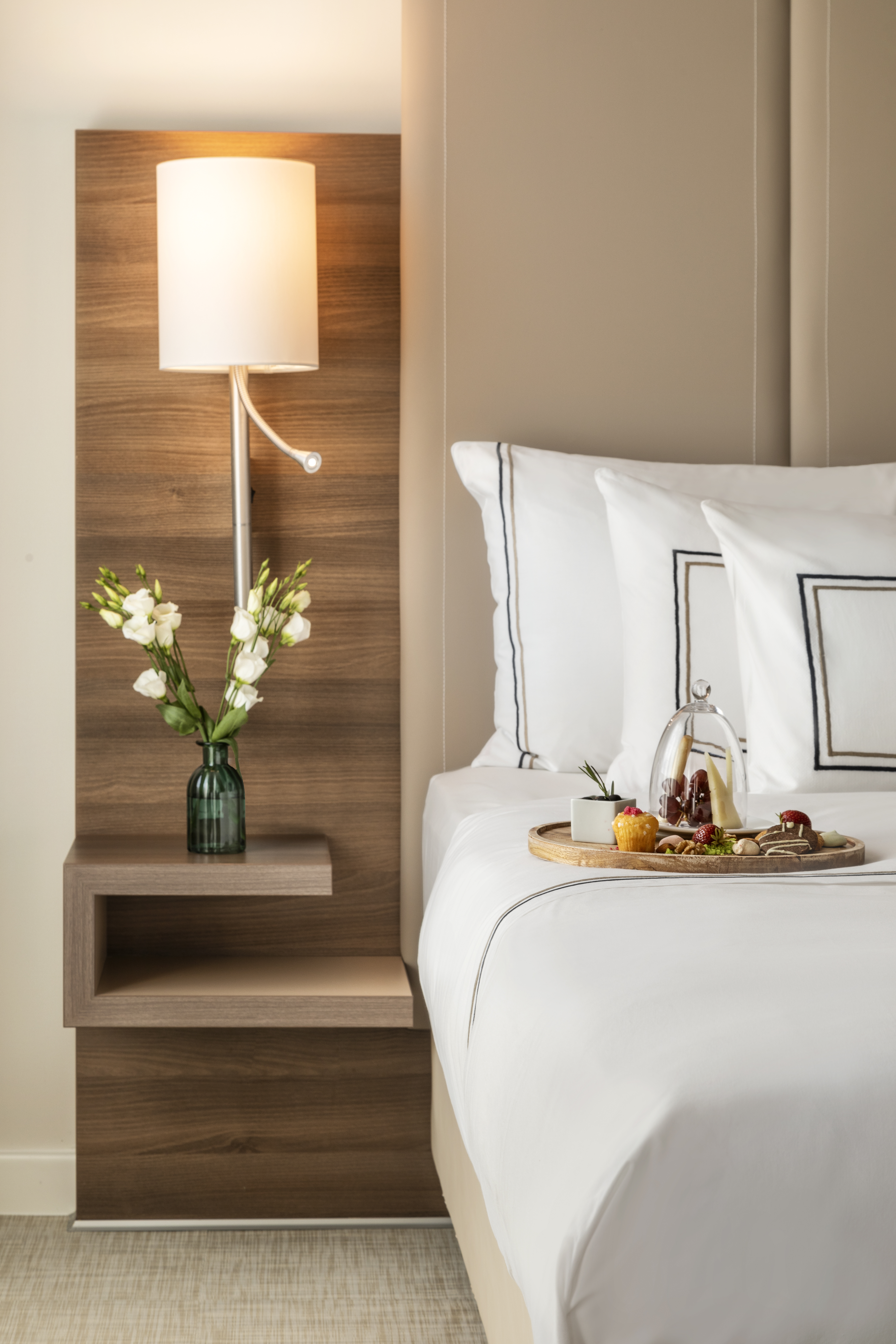 Hotel bed with snack tray, white linens, bedside lamp, and wooden nightstand.