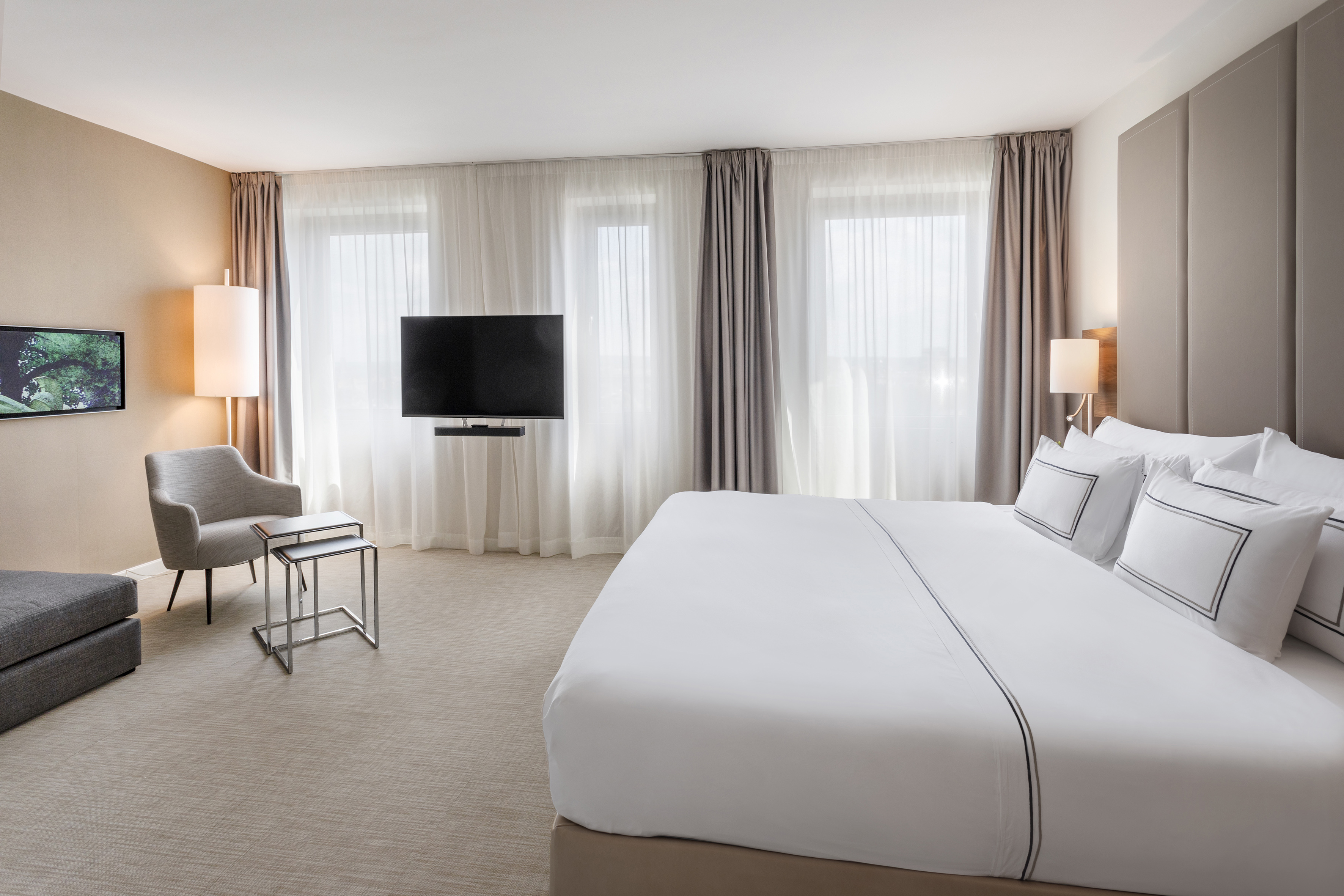 Modern hotel bedroom with king bed, wall TV, armchair, and bright windows.