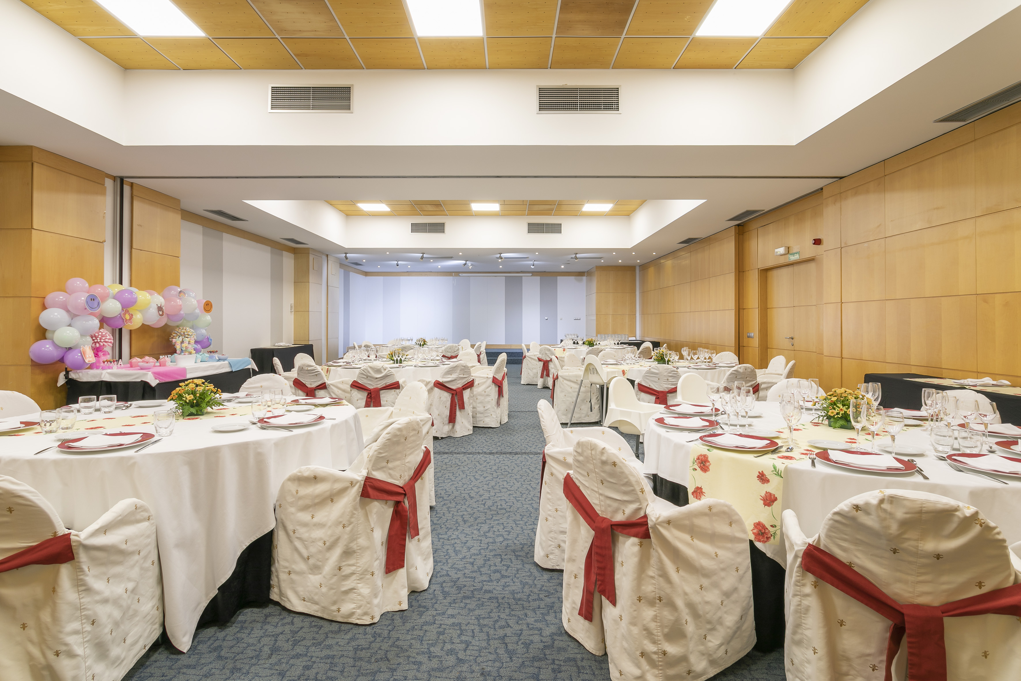 Banquet hall set for a party with white tables, red chair sashes, and balloons.