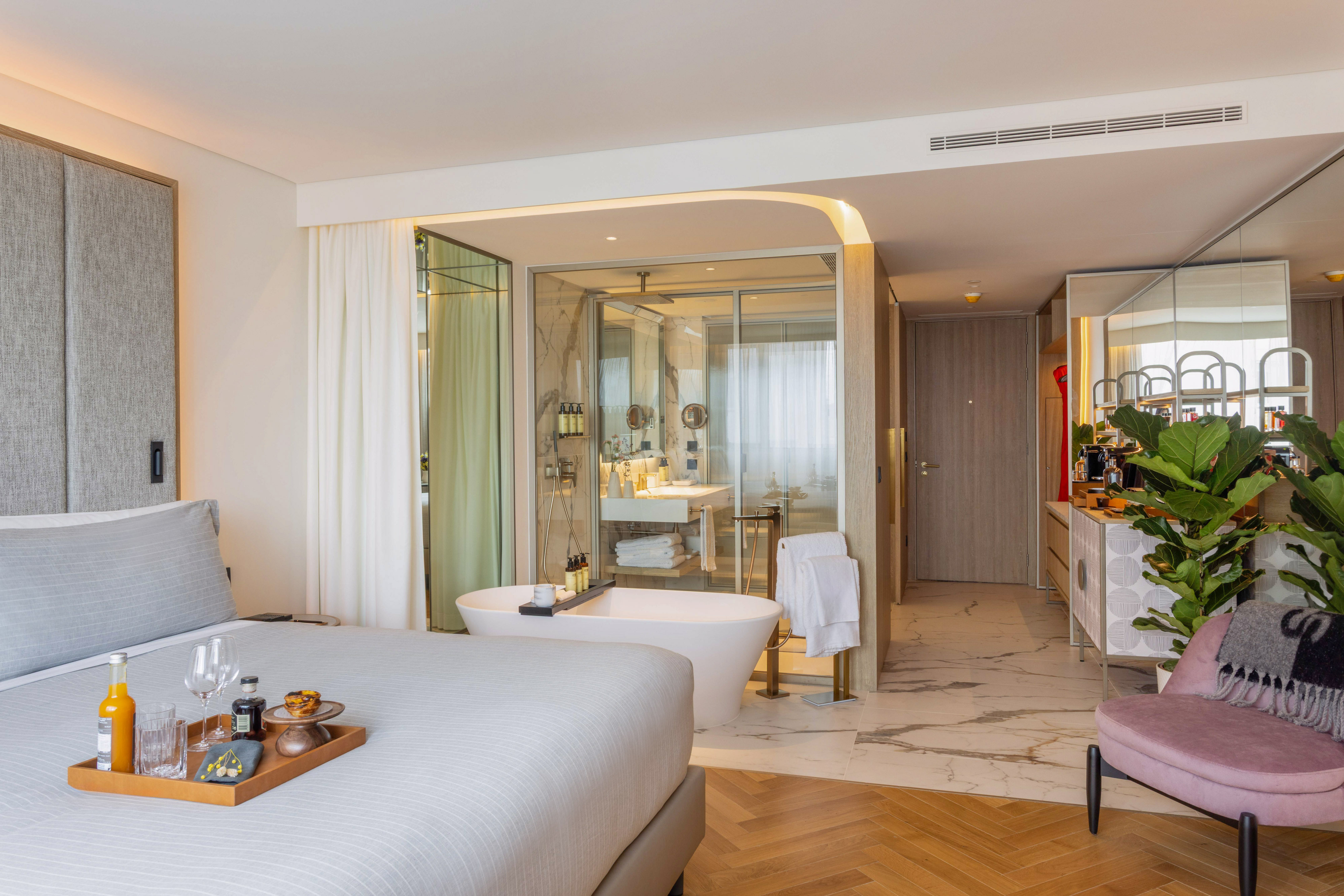 Modern hotel room with breakfast on bed, freestanding tub, and open bathroom.