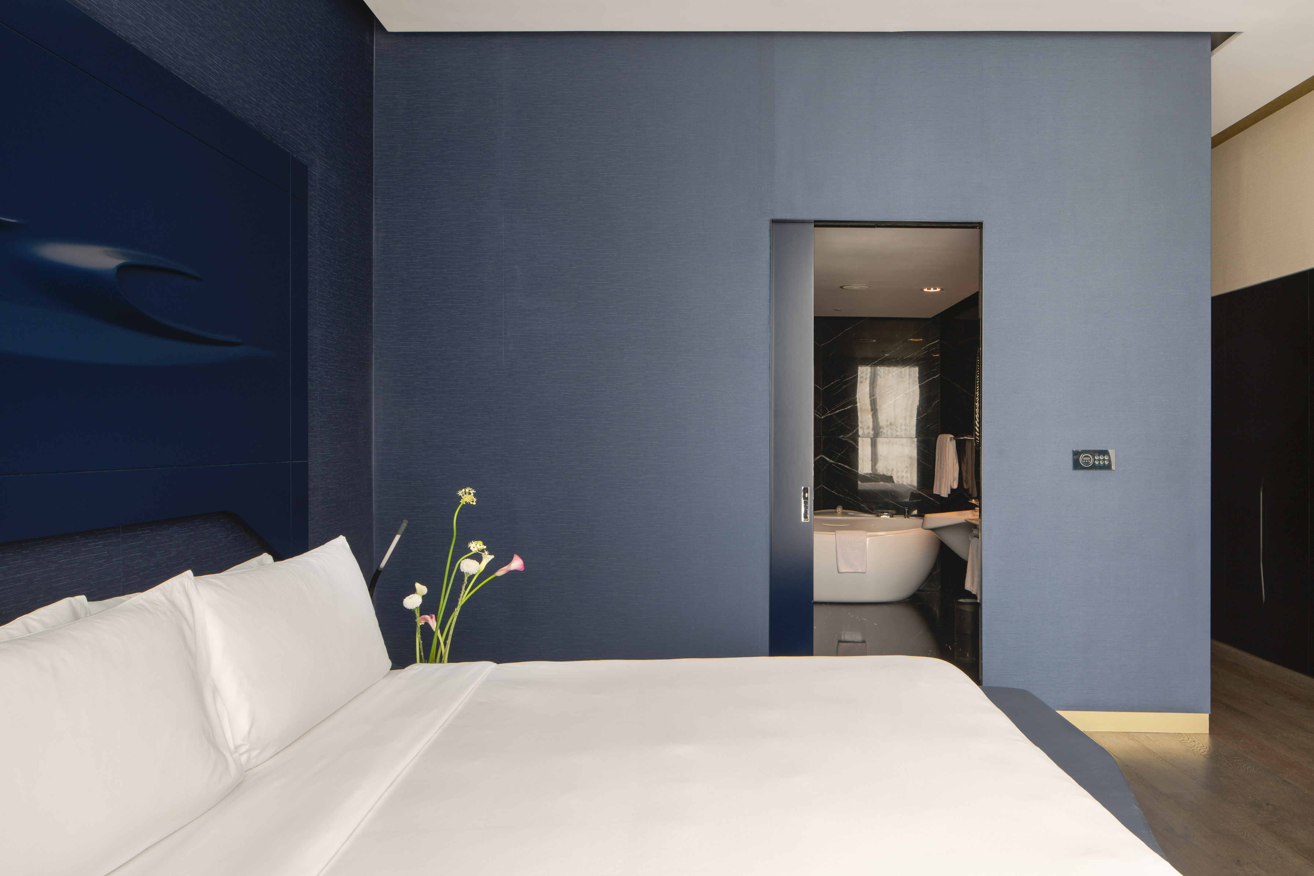 Modern luxury hotel room with white bed, navy walls, and open marble bathroom.
