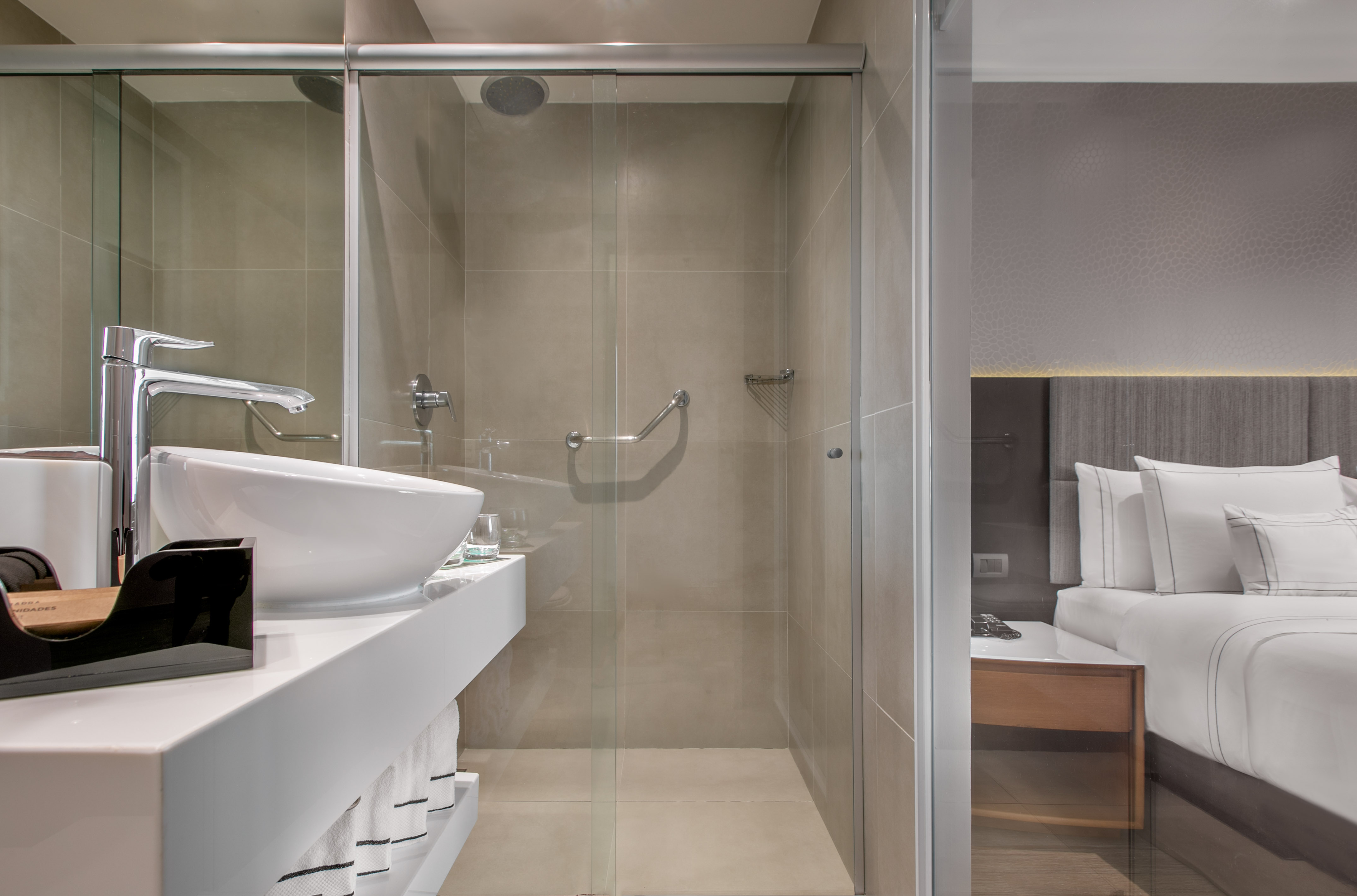 Sleek hotel bathroom with glass shower, white sink, and adjoining bedroom view.