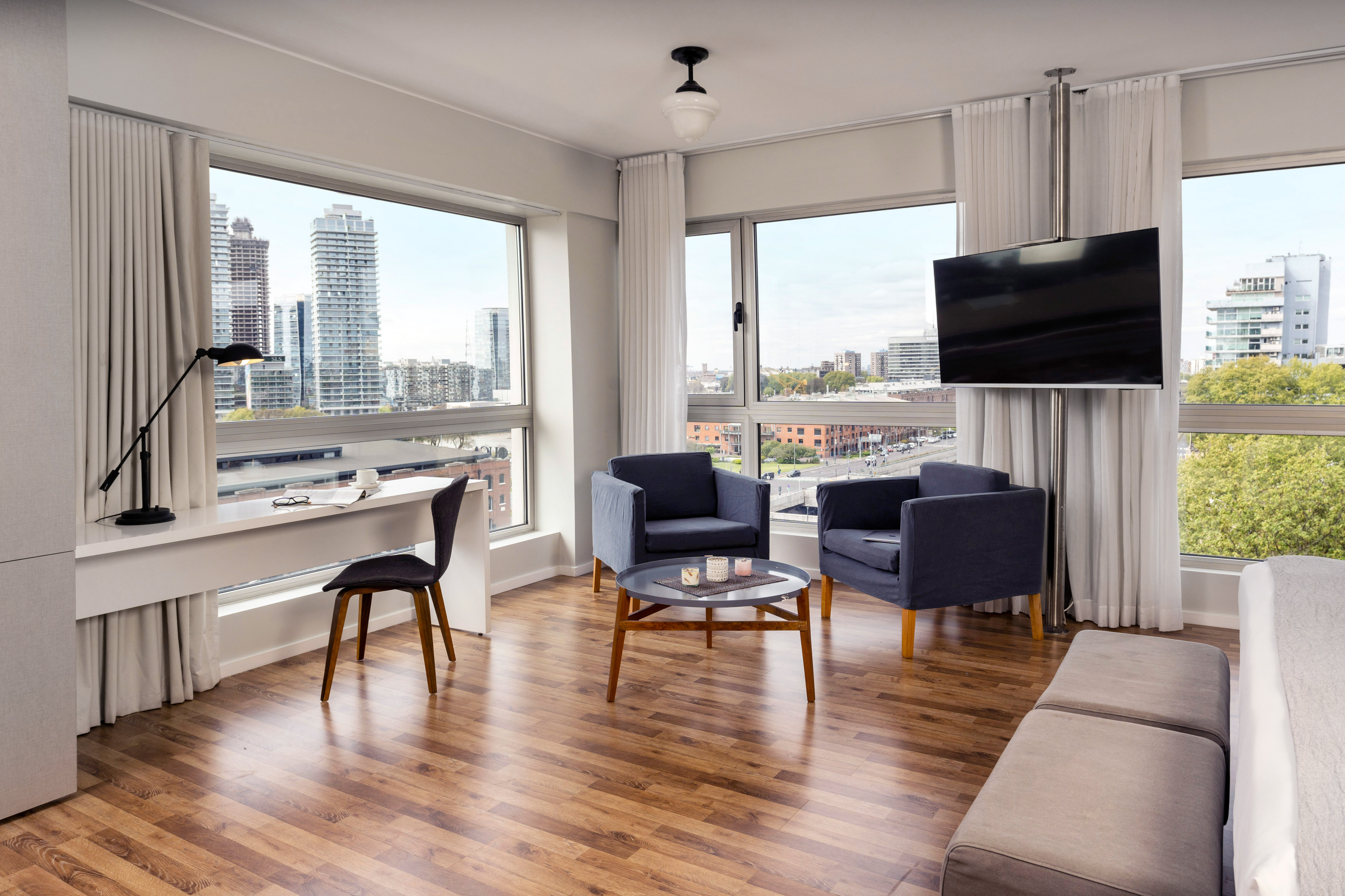 Modern hotel suite living room with city view, desk, armchairs, and TV.