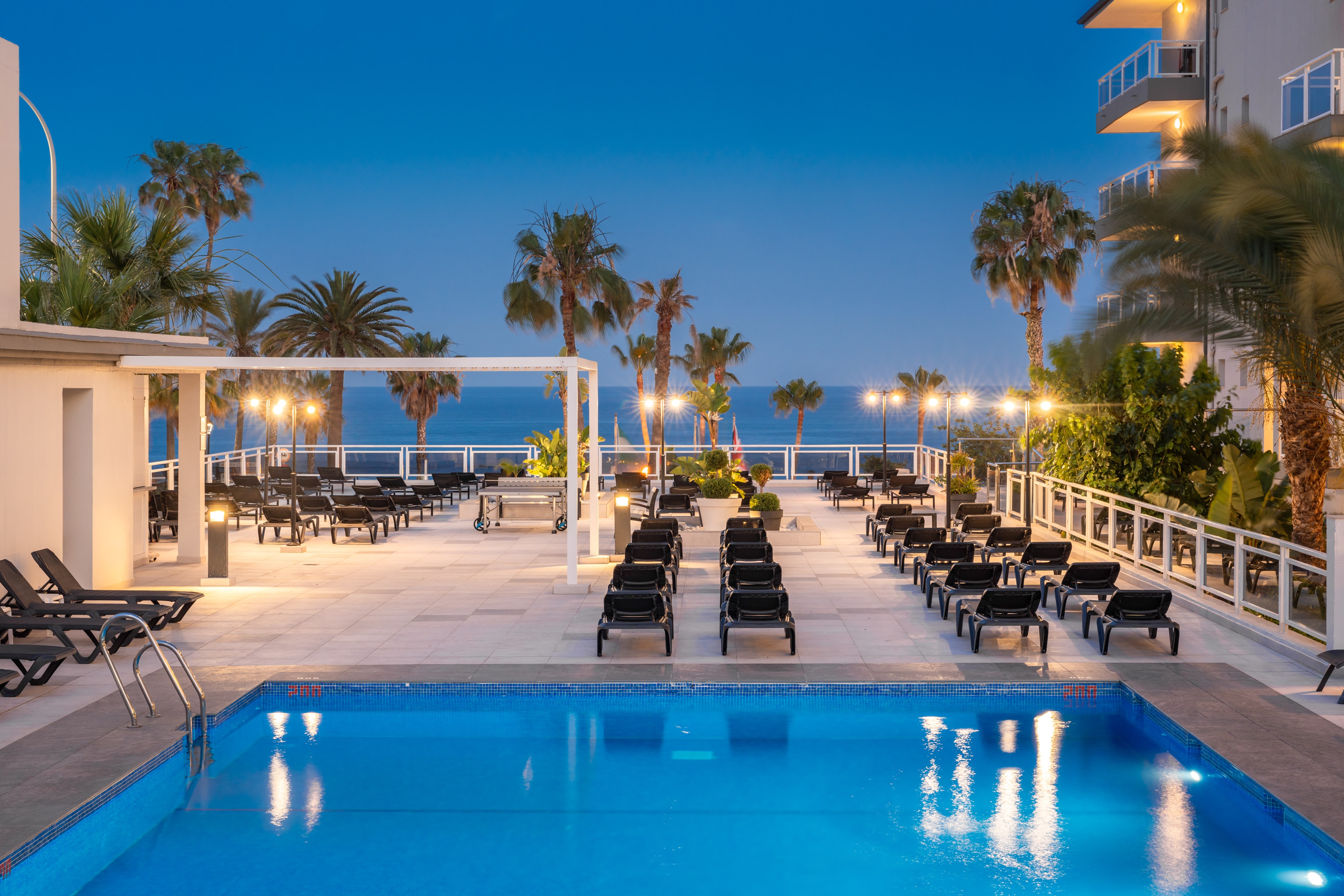 Resort swimming pool, sun loungers, palm trees, and ocean view at dusk.