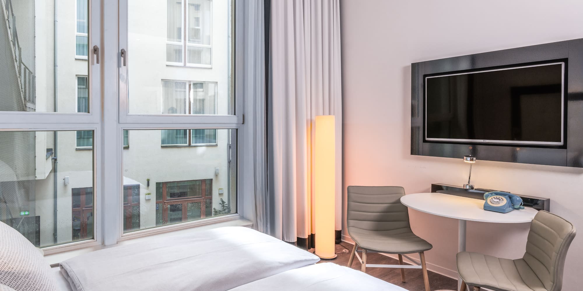 Modern hotel room with bed, window, wall TV, small table, and retro phone.