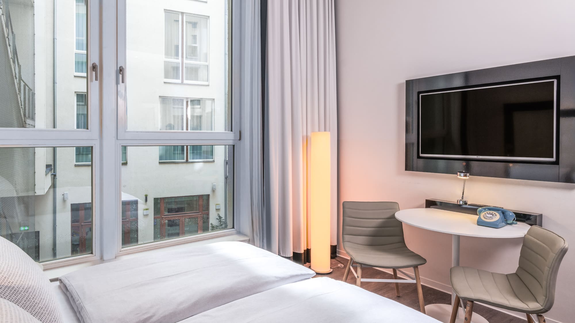 Modern hotel room with bed, window, wall TV, small table, and retro phone.