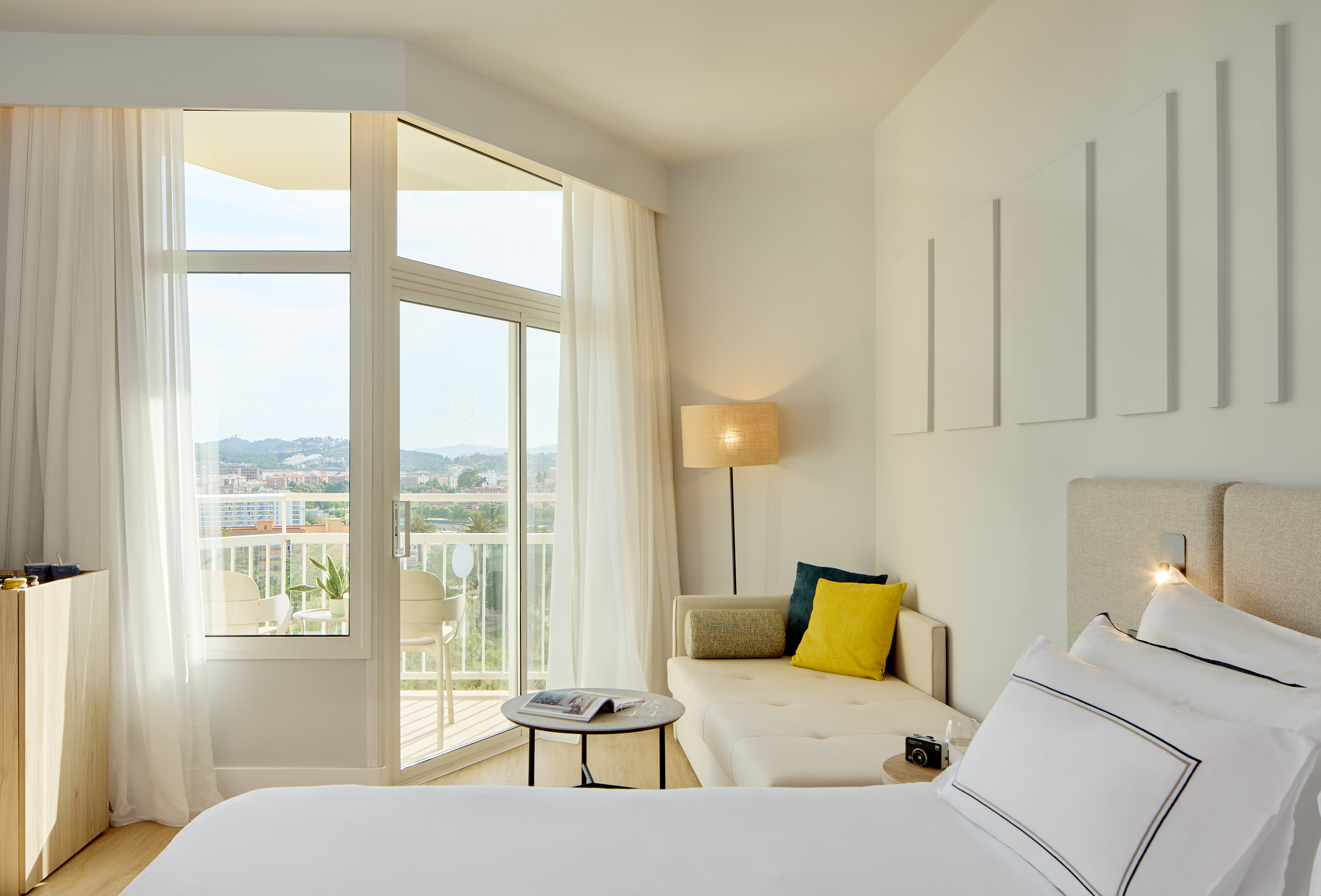 Bright modern hotel room with bed, sofa, balcony, and urban view.