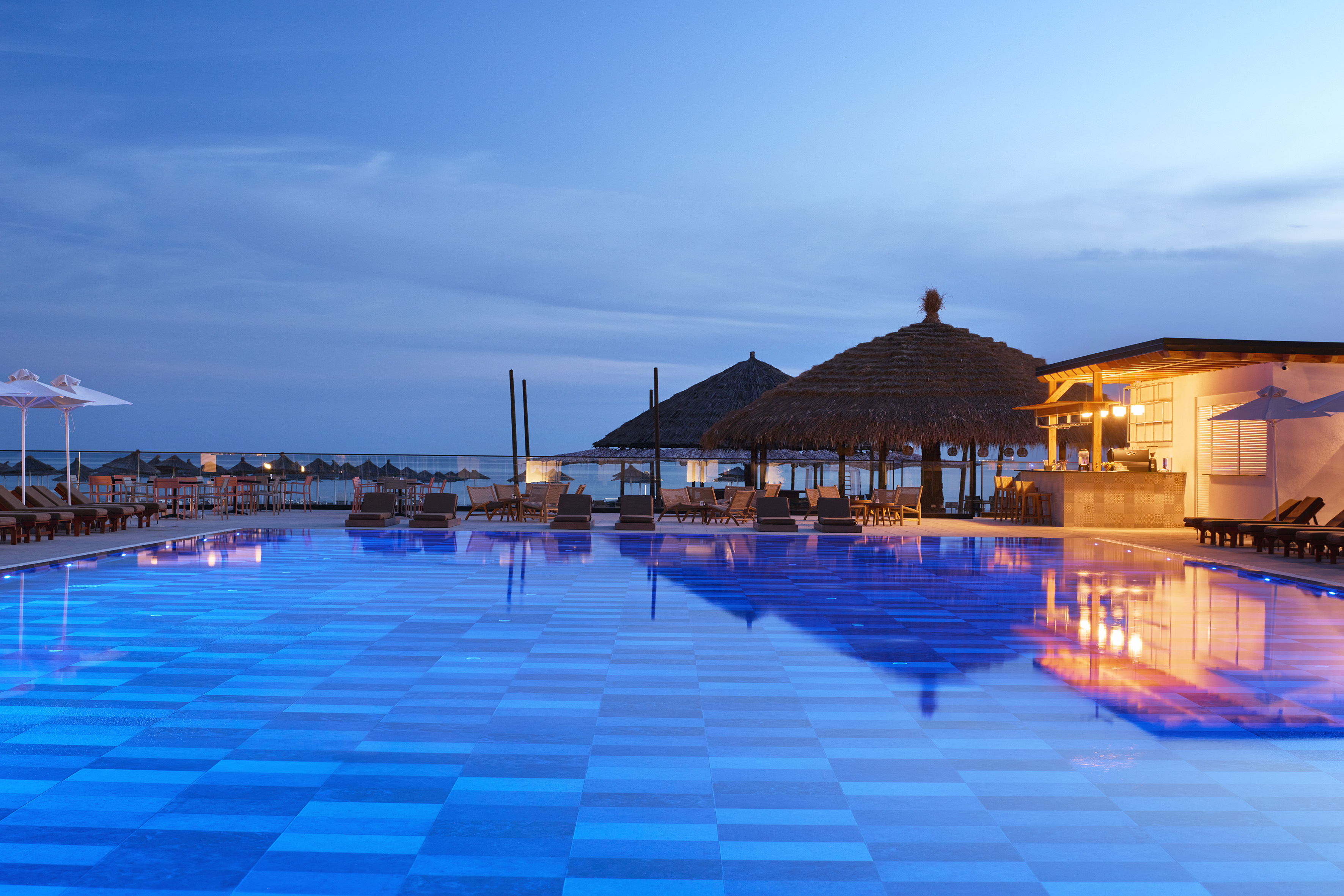 Luxury resort pool, illuminated blue tiles, thatched bar at dusk.