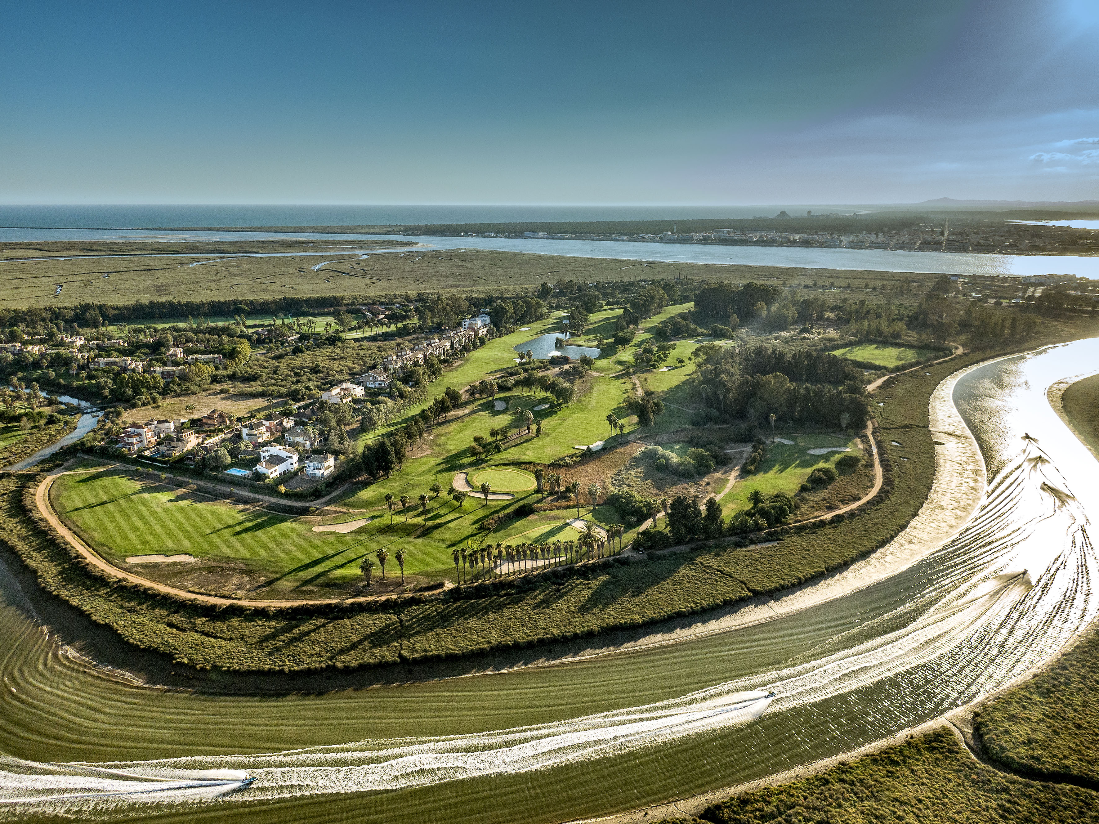 Drone view of coastal golf course with river, speedboats, and residential area.