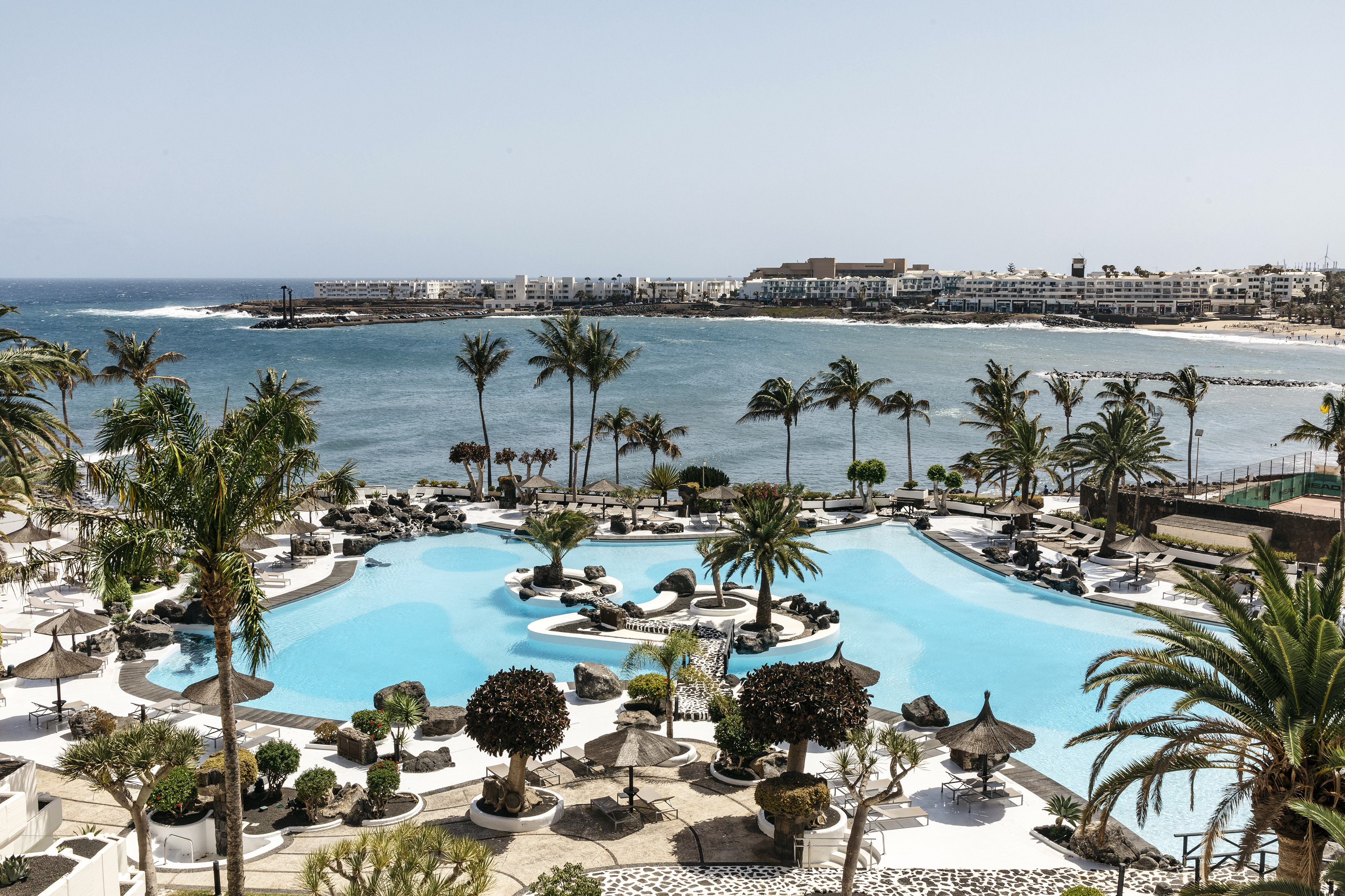 Resort lagoon pool with palm trees, ocean view, and coastal buildings.